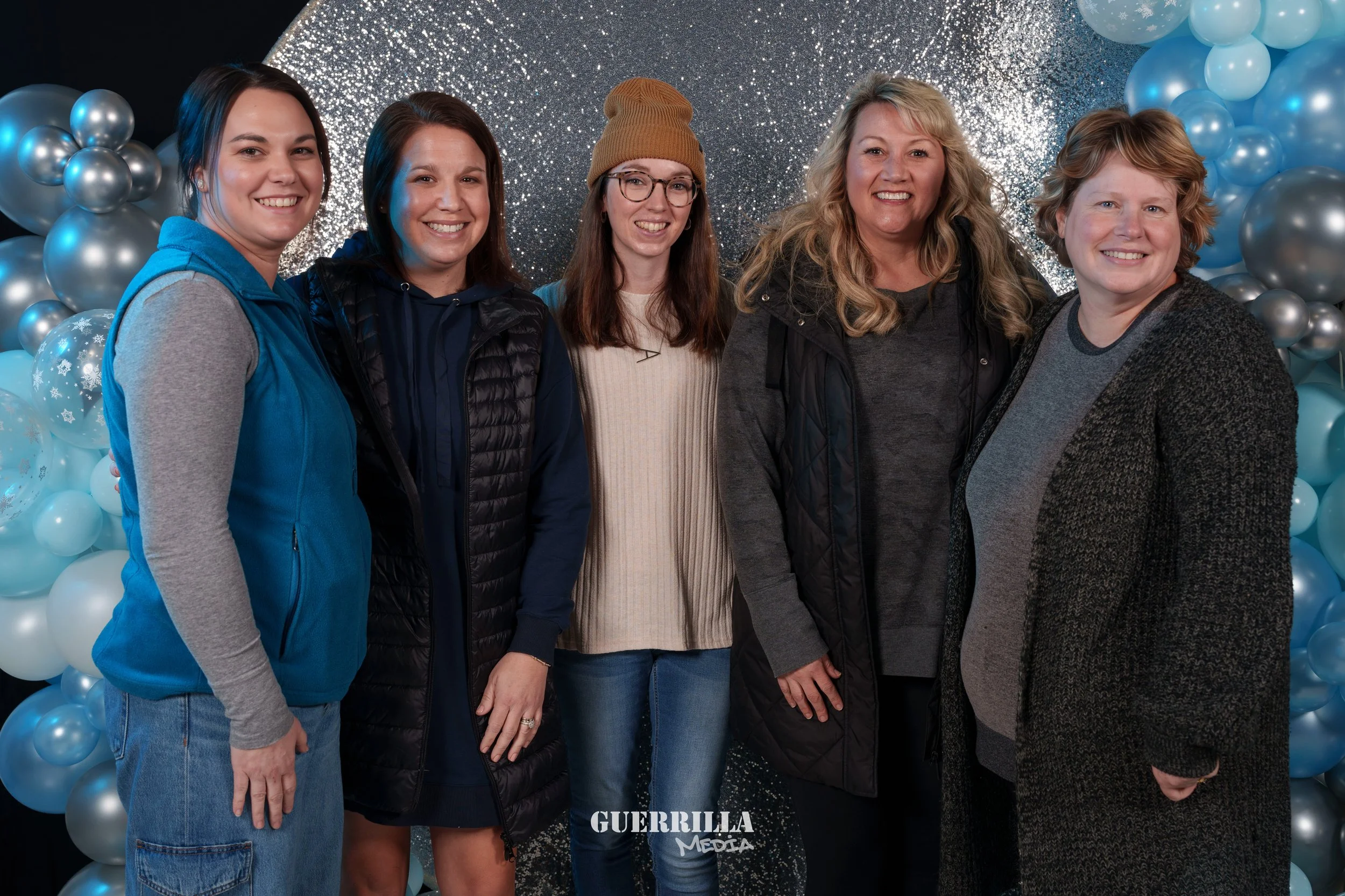 Group of five women standing together in front of a blue and silver balloon backdrop, smiling at the camera.
