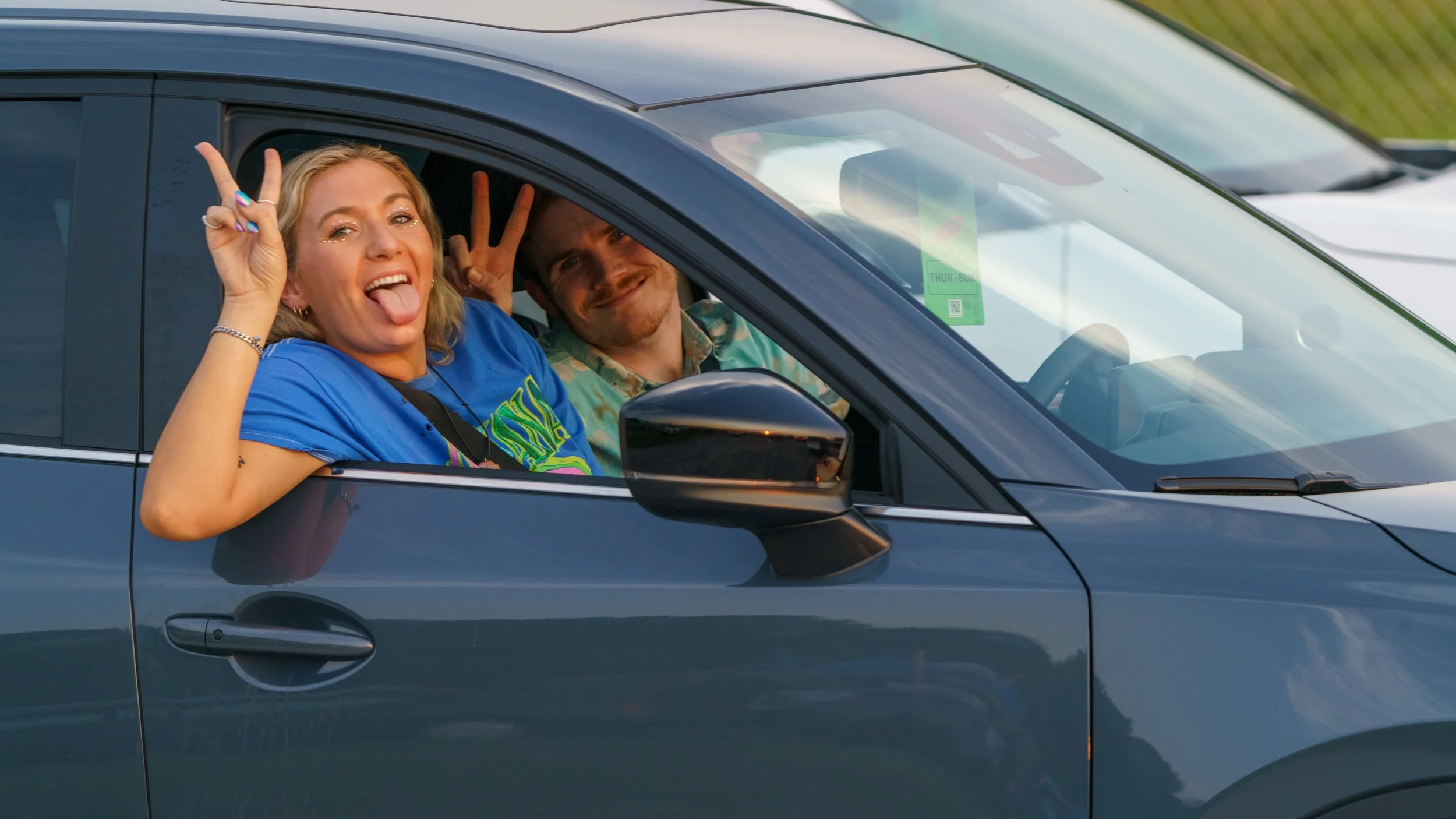 Two young adults in a car, woman in the front seat making a peace sign and sticking out her tongue, and man in the back seat making a peace sign, both smiling.