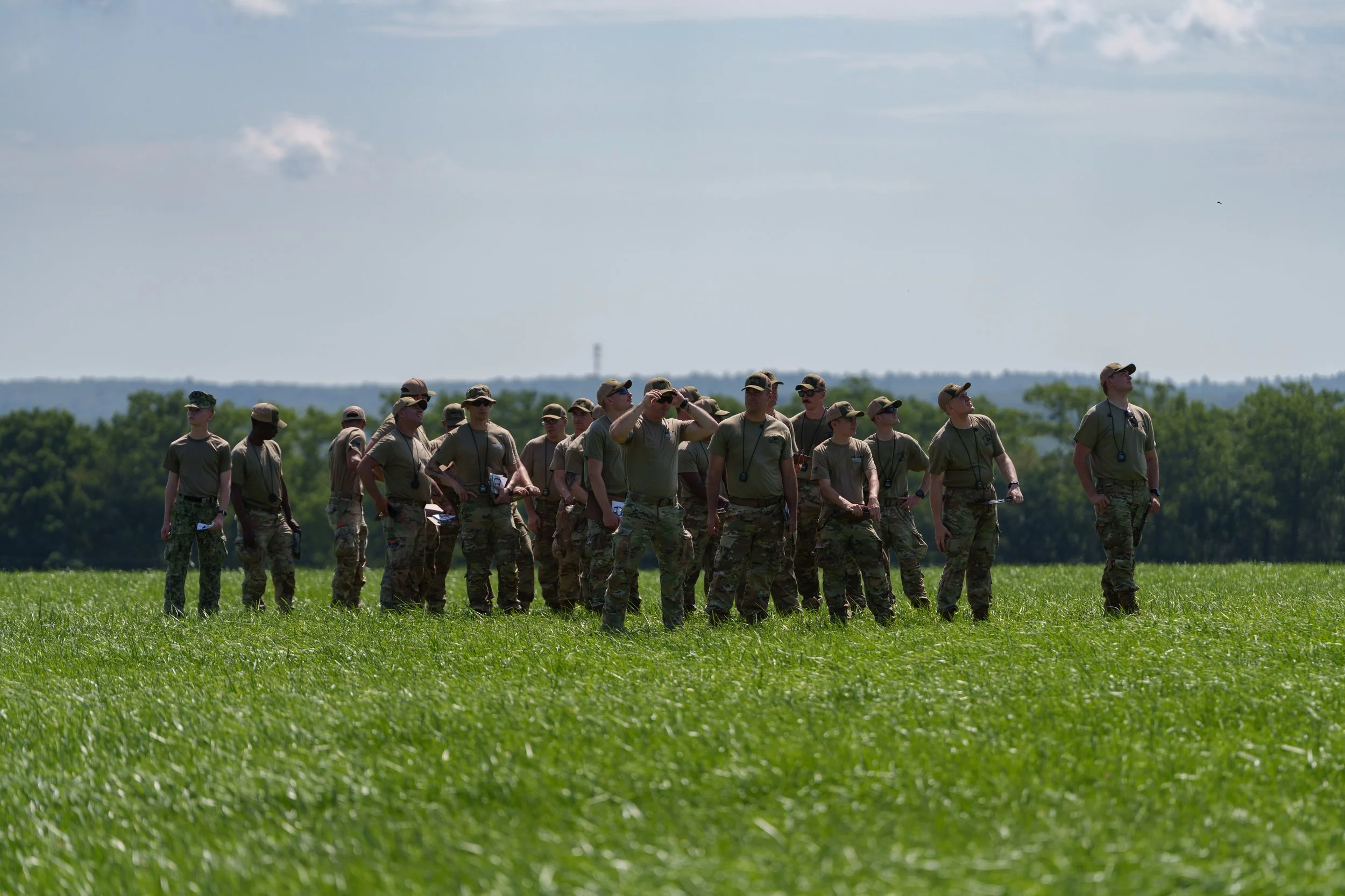 A group of soldiers in military uniforms standing in a grassy field, some looking around and others using binoculars or cameras, with a background of trees and a partly cloudy sky.
