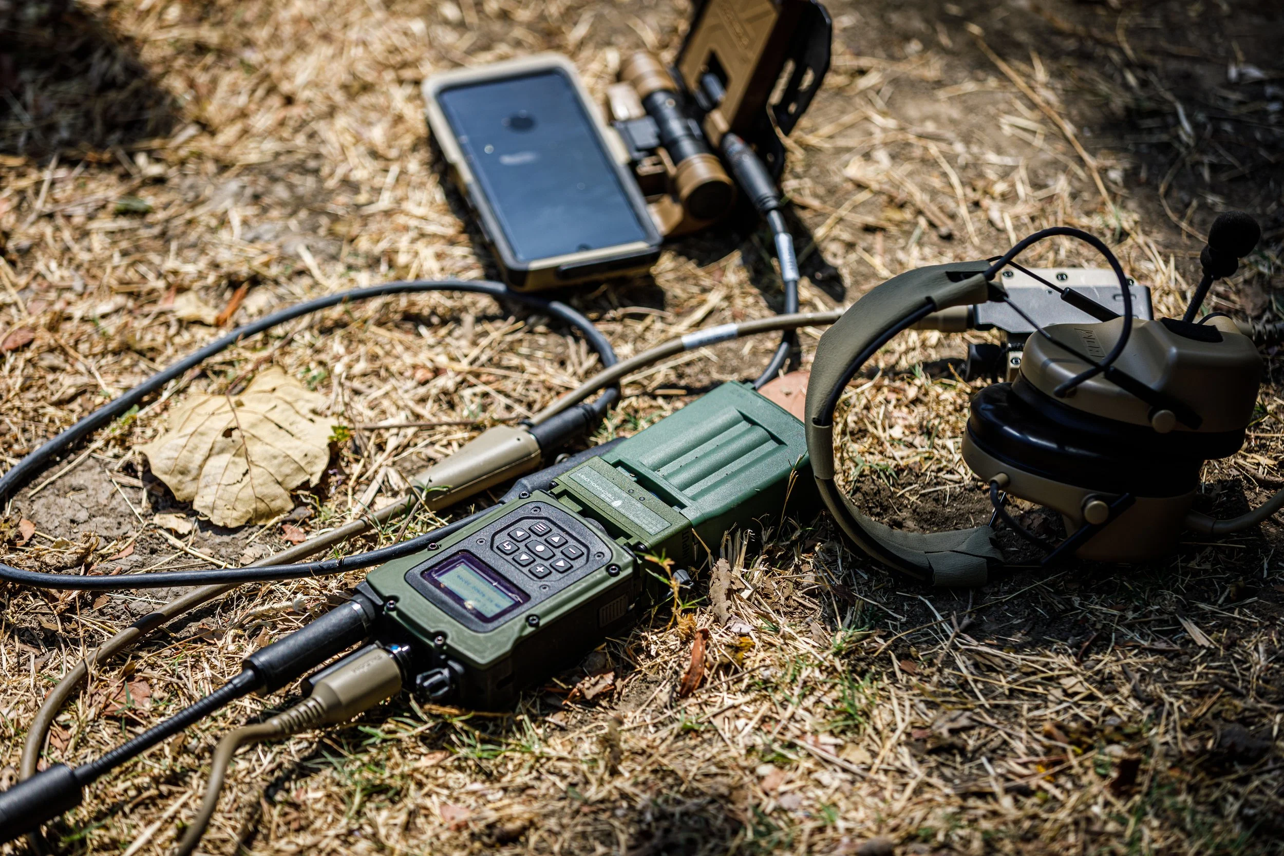 Metal detector, headphones, and a smartphone on dry grass, with the headphones resting on the ground and the metal detector turned on.