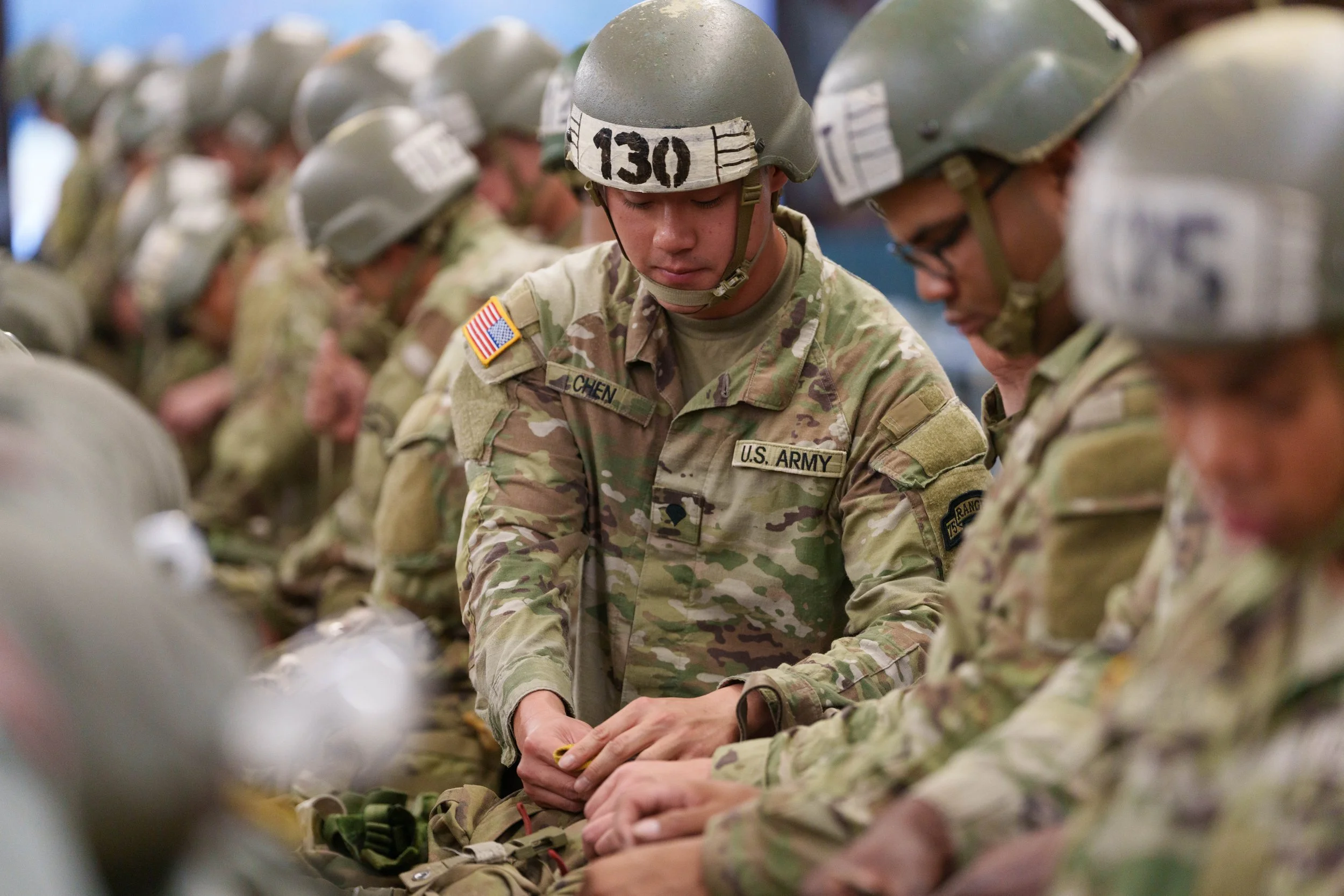 U.S. soldiers in camouflage uniforms and helmets standing in a row with their heads bowed, appearing to be in a moment of prayer or reflection.
