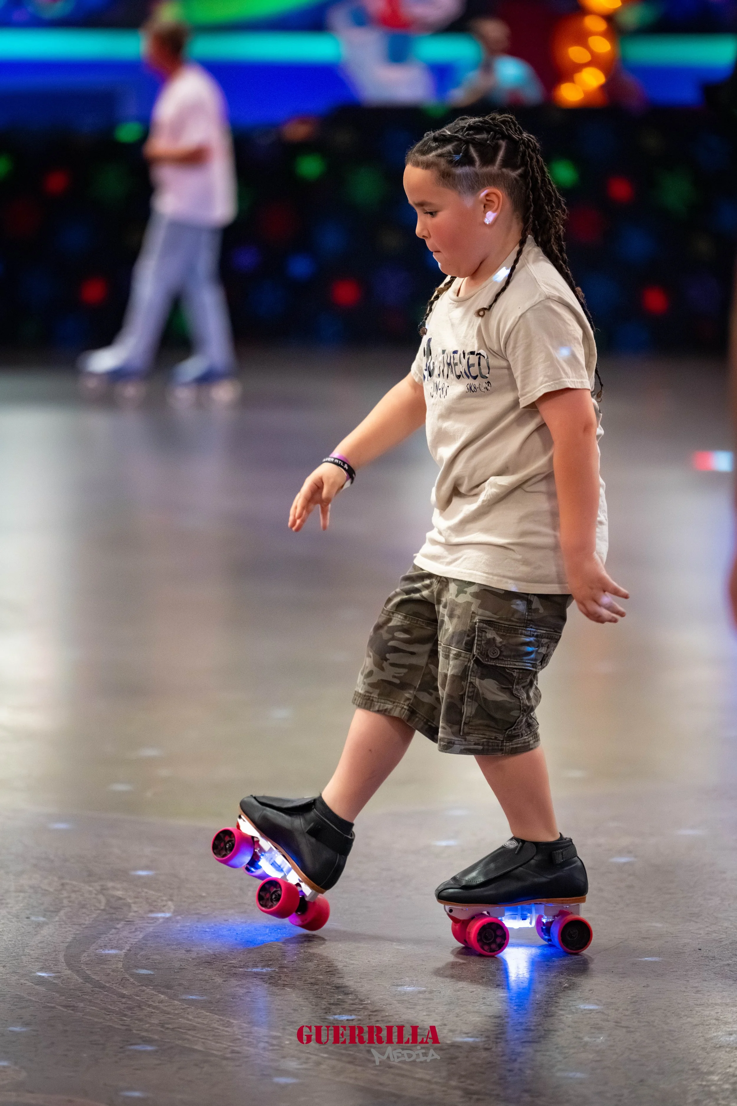 A young girl with braided hair roller skating indoors, with a colorful blurred background and another person in the distance.