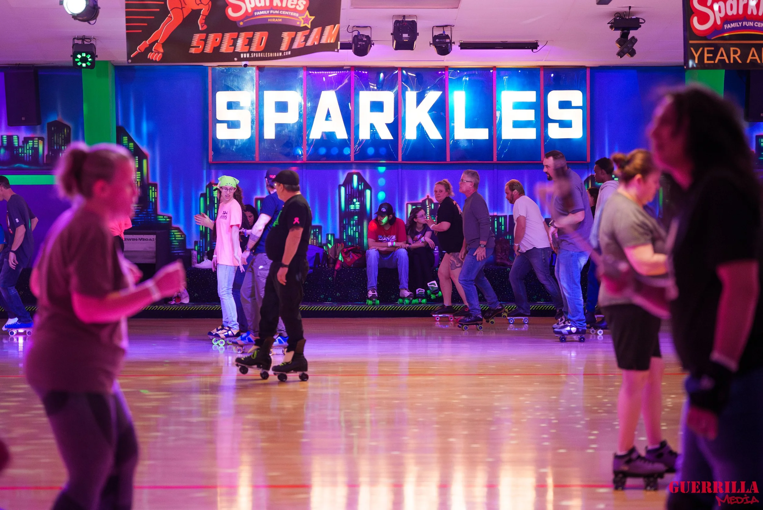 People roller skating inside an indoor skating rink with colorful neon lights and a large illuminated sign that says 'SPARKLES' in the background.