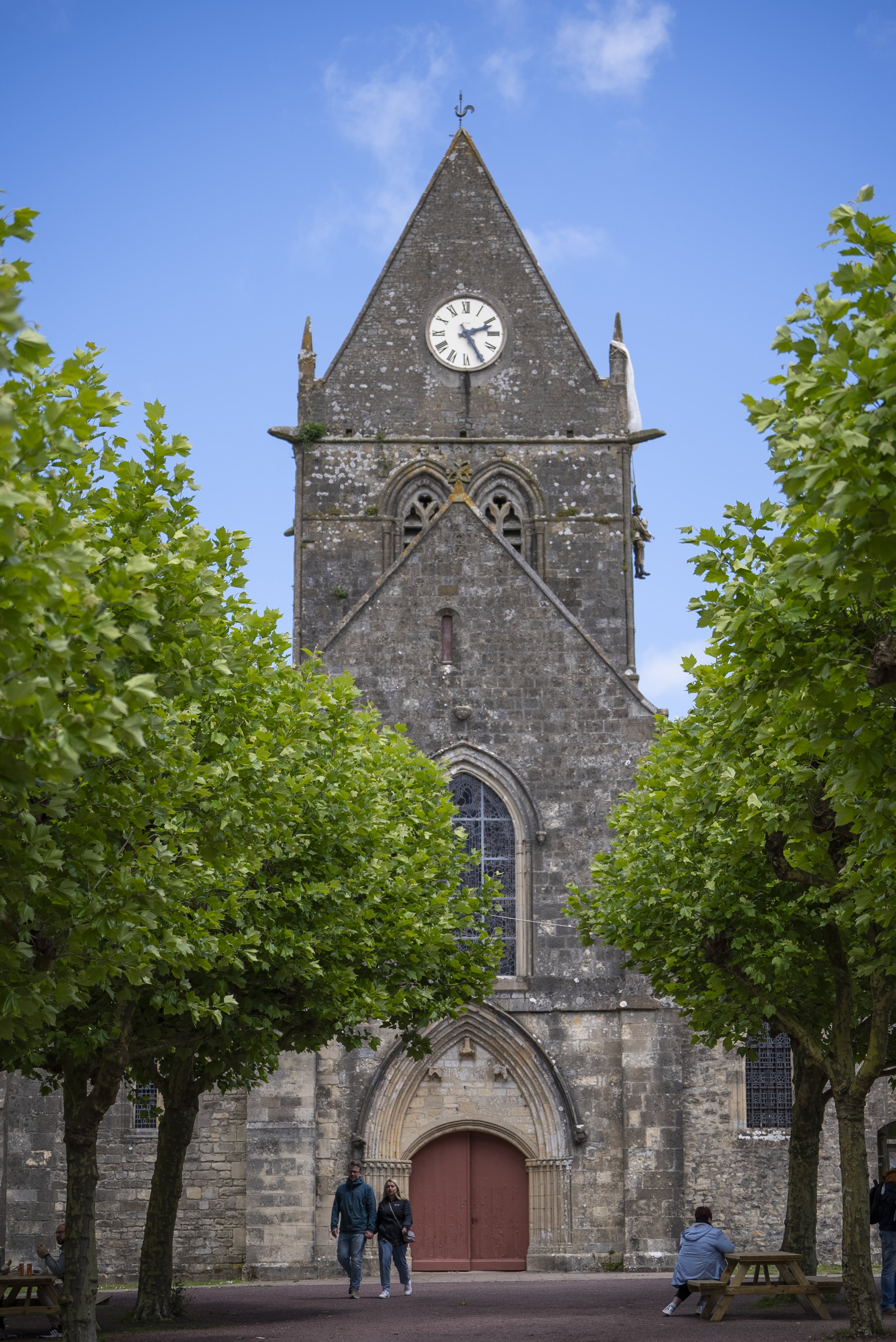 A historic stone church with a clock tower, surrounded by green leafy trees, with people walking and sitting nearby on a paved area.