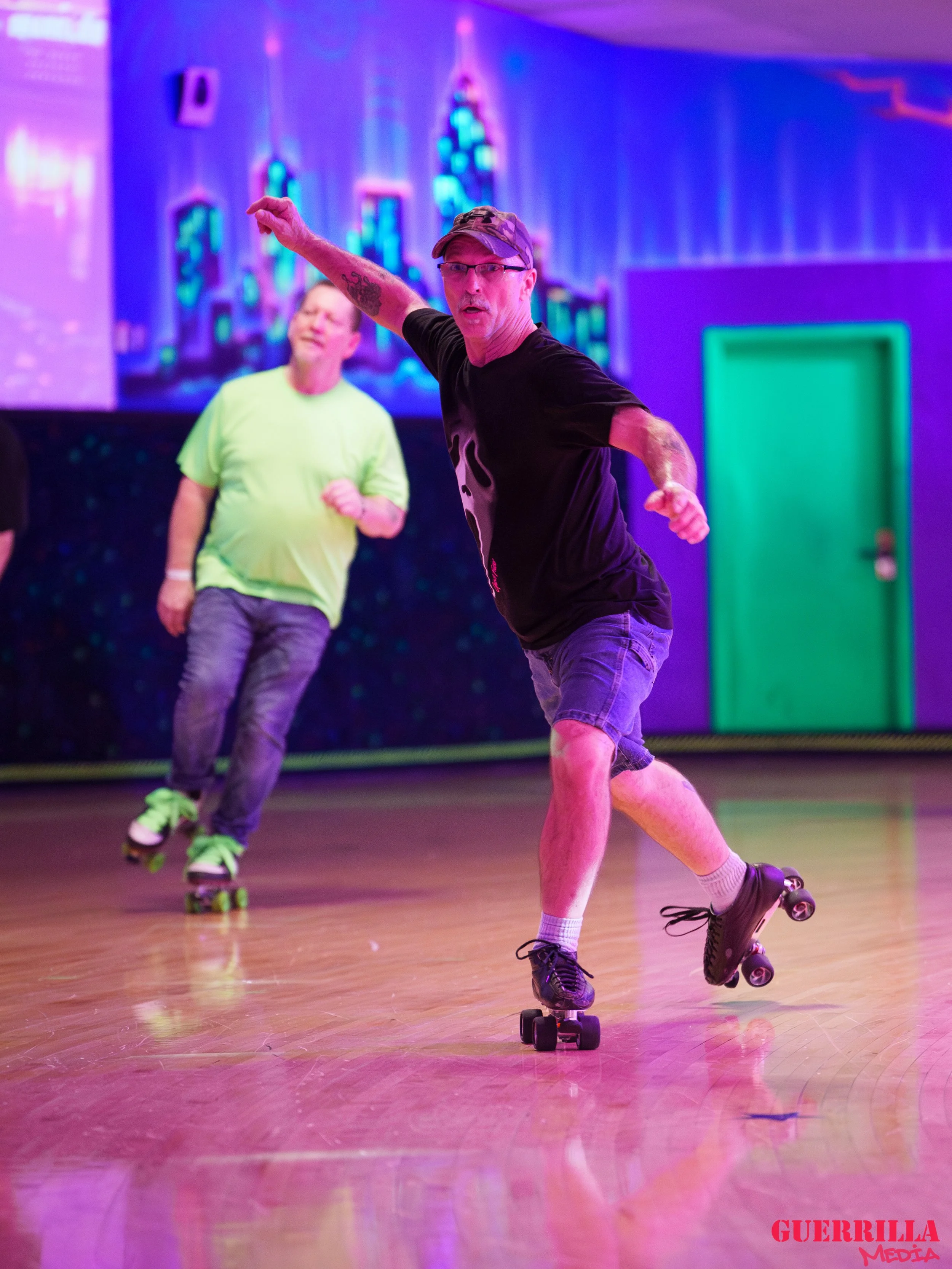 Two men roller skating indoors under vibrant purple and green lighting with a cityscape digital background.