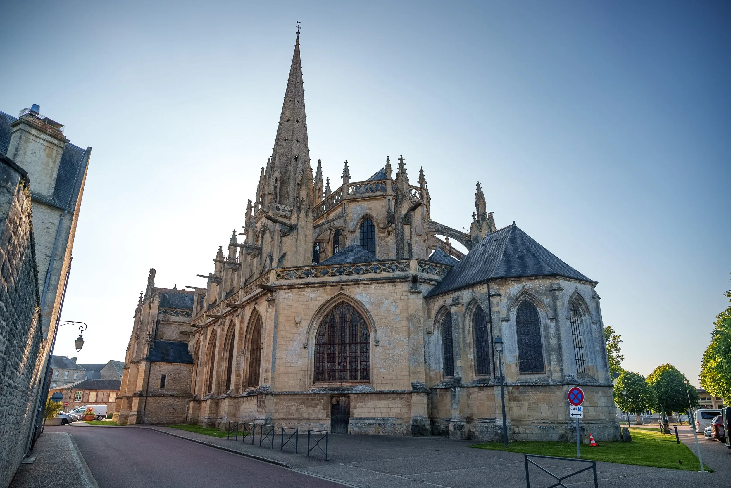 A historic Gothic church with tall spire, large stained glass windows, and stone architecture, set in a small town with trees and parked cars.