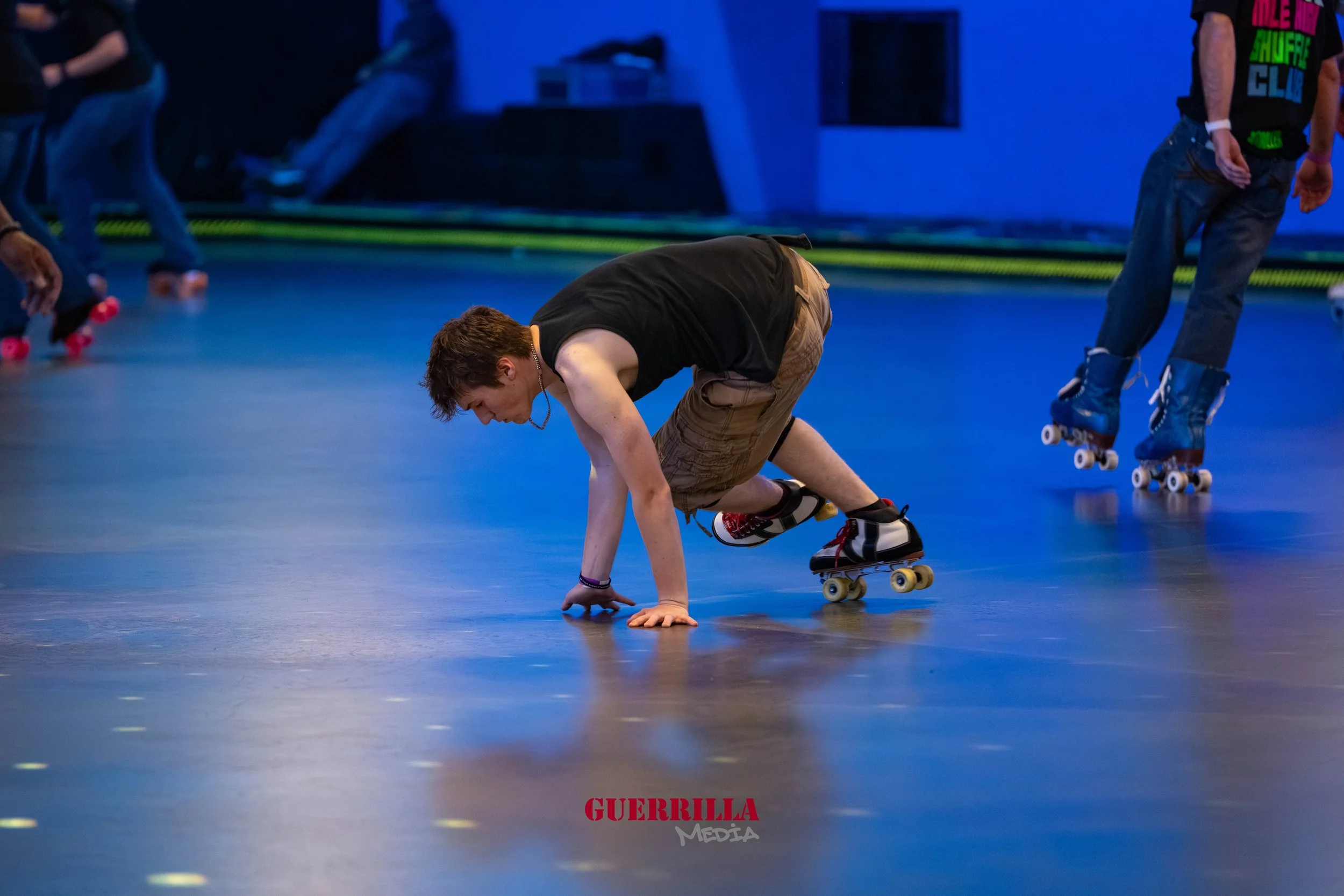 Young man in a black tank top and brown shorts crouching on roller skates inside a roller skating rink, with other skaters in the background.