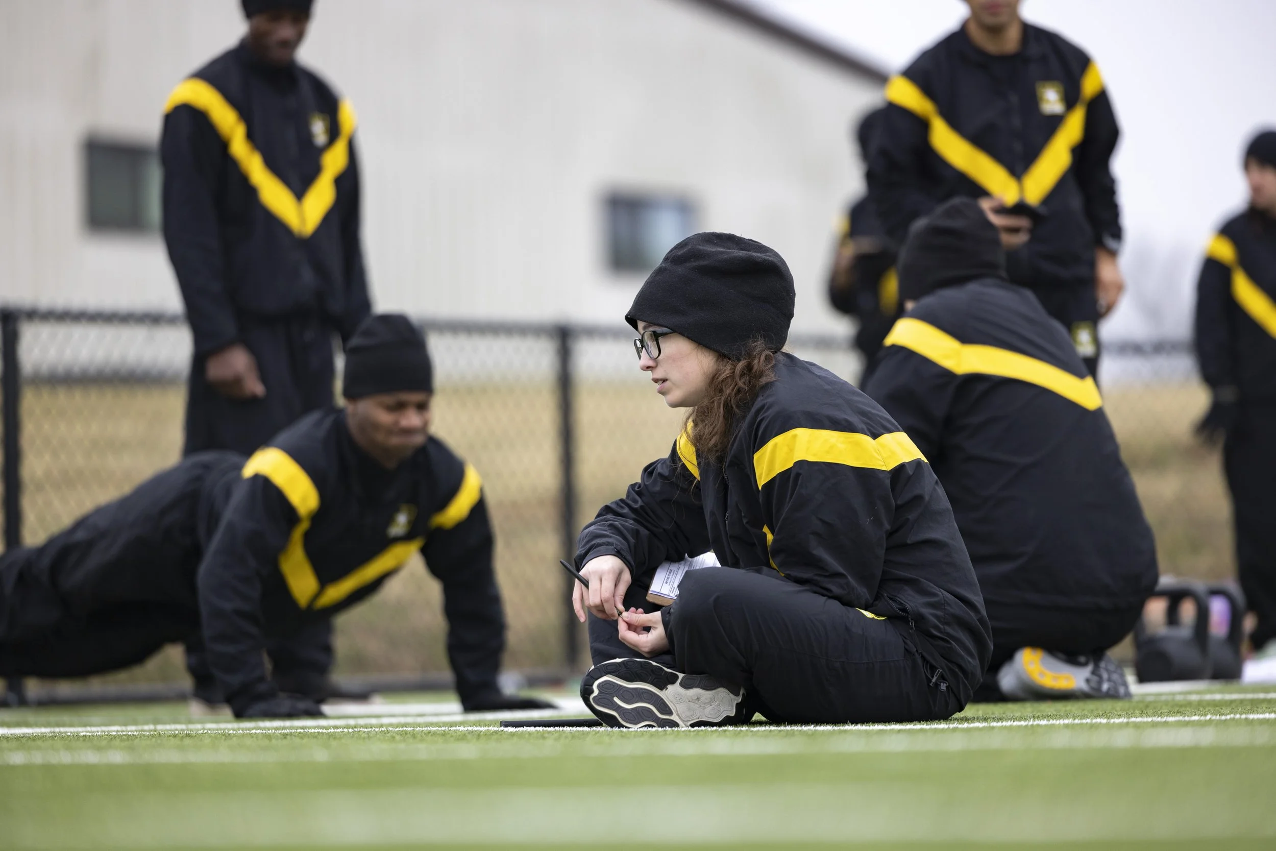 A group of athletes in black jackets with yellow stripes, some wearing beanies, participating in an outdoor workout. One woman is sitting on the ground holding a pen, and others are in push-up position or standing nearby, on a sports field with a fen