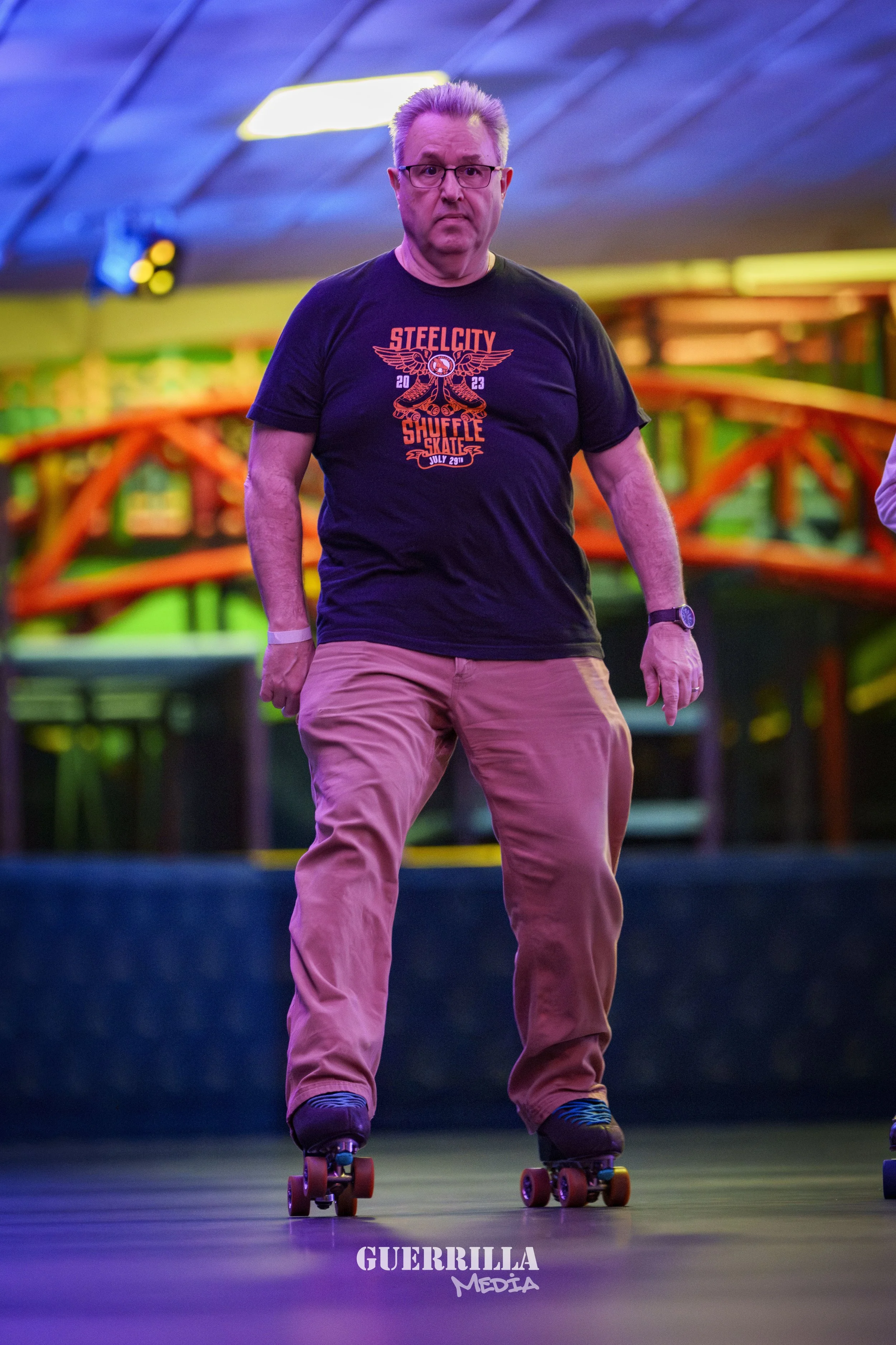 A man with glasses and gray hair roller skating indoors with colorful lighting and a blurred background.