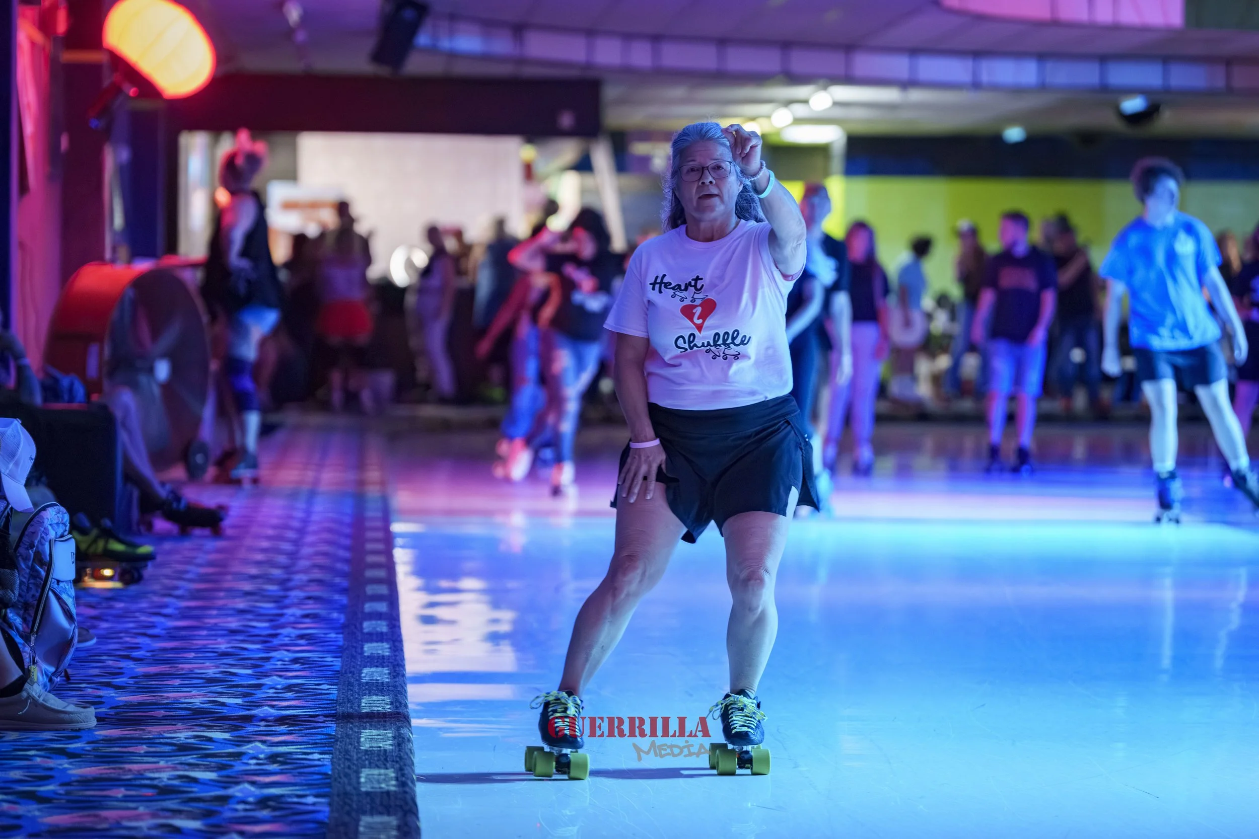 An older woman roller skating indoors at a roller rink with colorful lights, wearing a white t-shirt and black shorts, with people in the background.