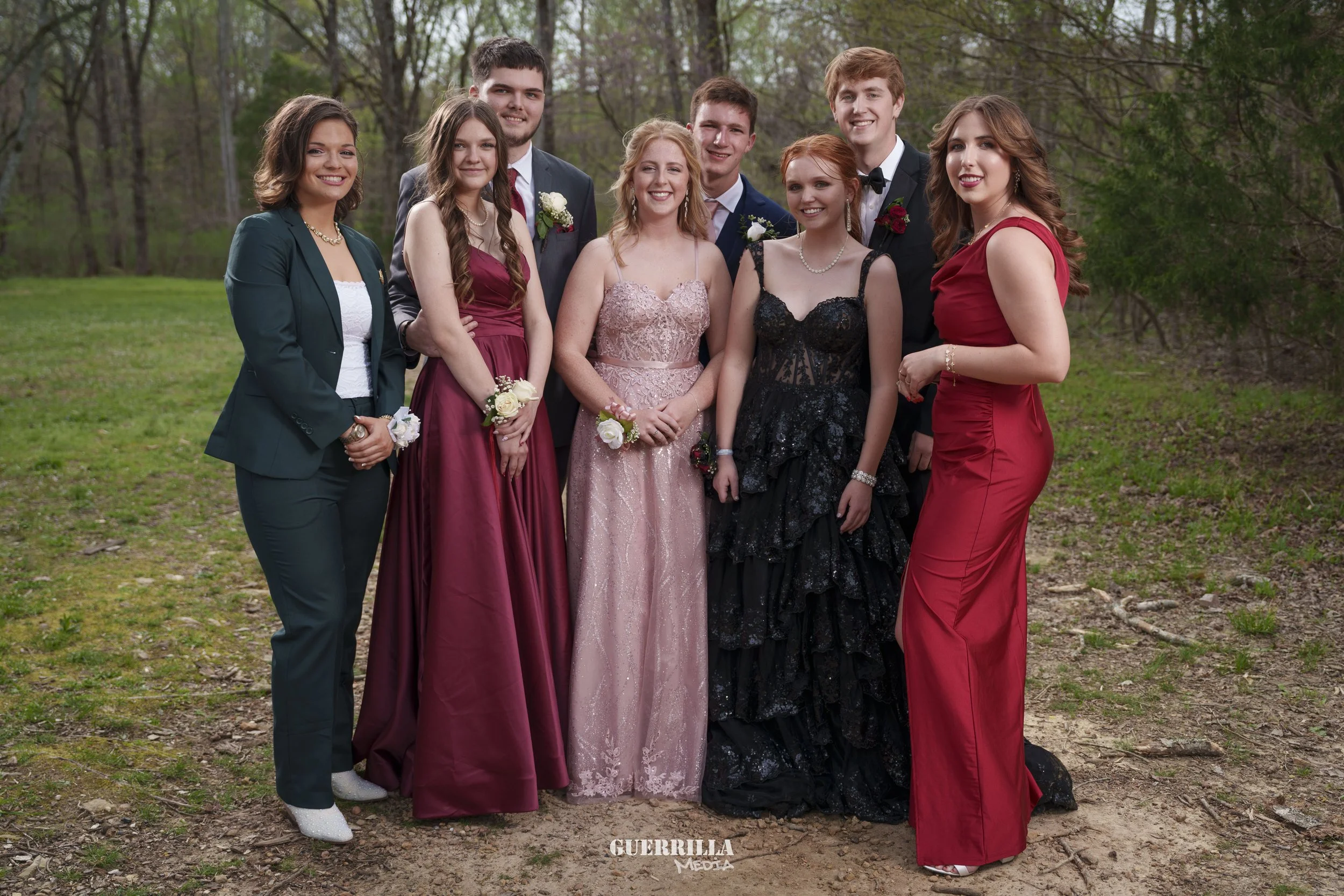 Group of nine teenagers dressed in formal attire, standing on a grassy outdoor area with trees in the background, posing for a photo.