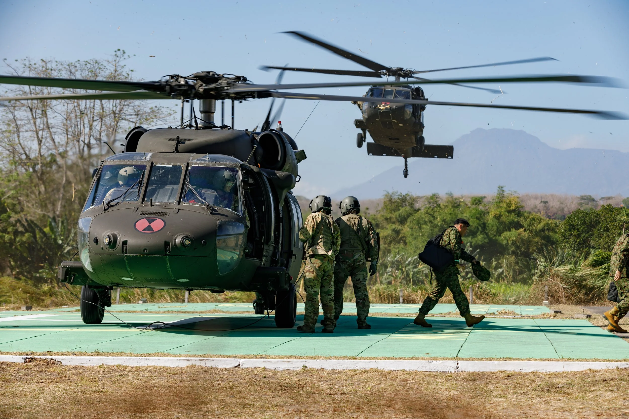 Military personnel boarding and preparing helicopters for flight in a natural landscape with mountains in the background.