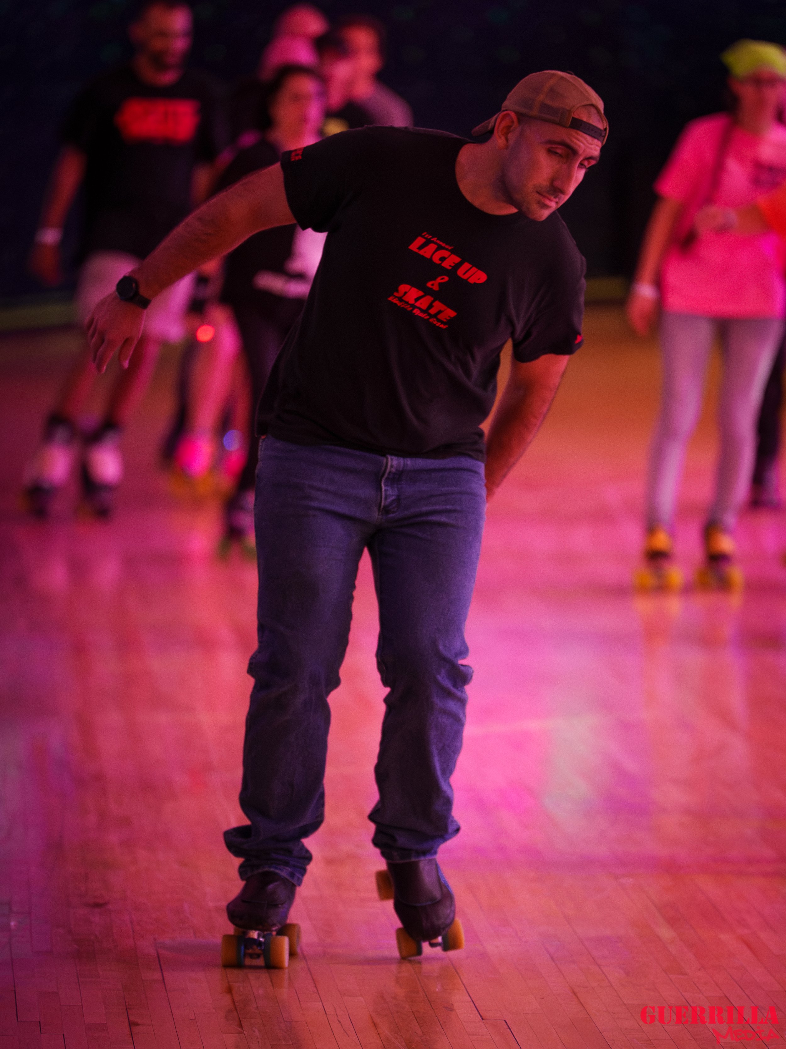 A man wearing a black T-shirt, blue jeans, and a backward cap is roller skating on a wooden floor. He is leaning forward with a serious expression, with a group of people in the background.