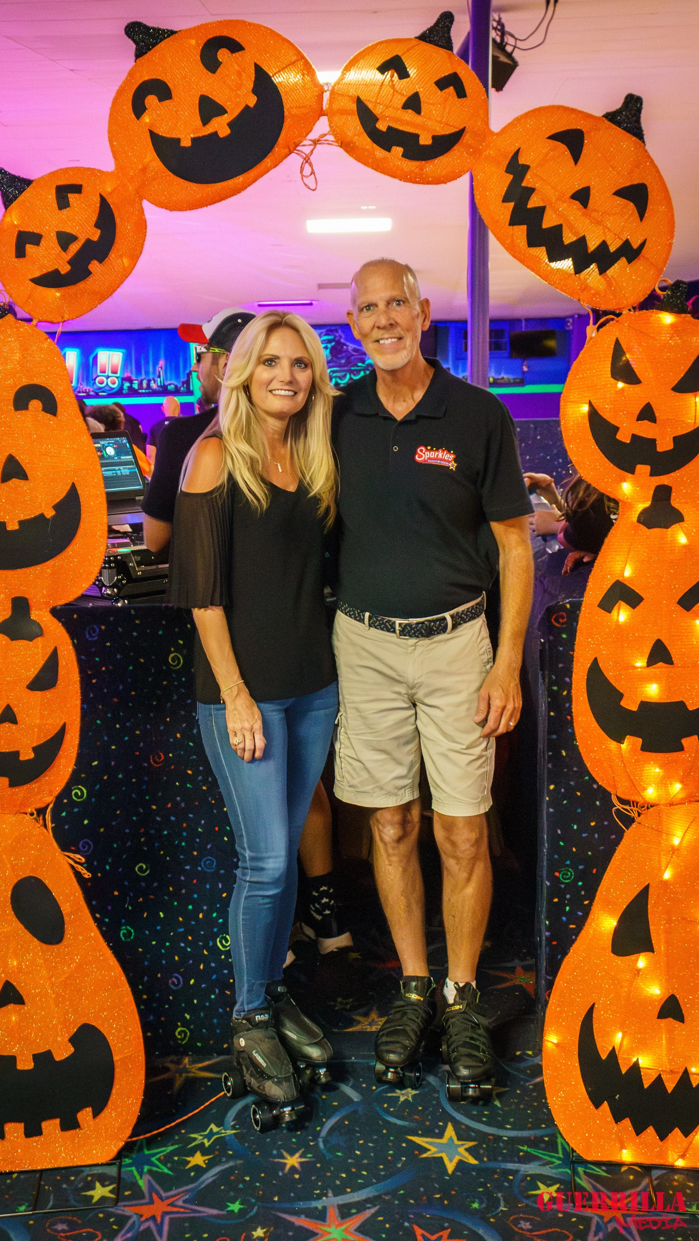 Two people wearing roller skates standing underneath a Halloween pumpkin decoration arch at a party or event.