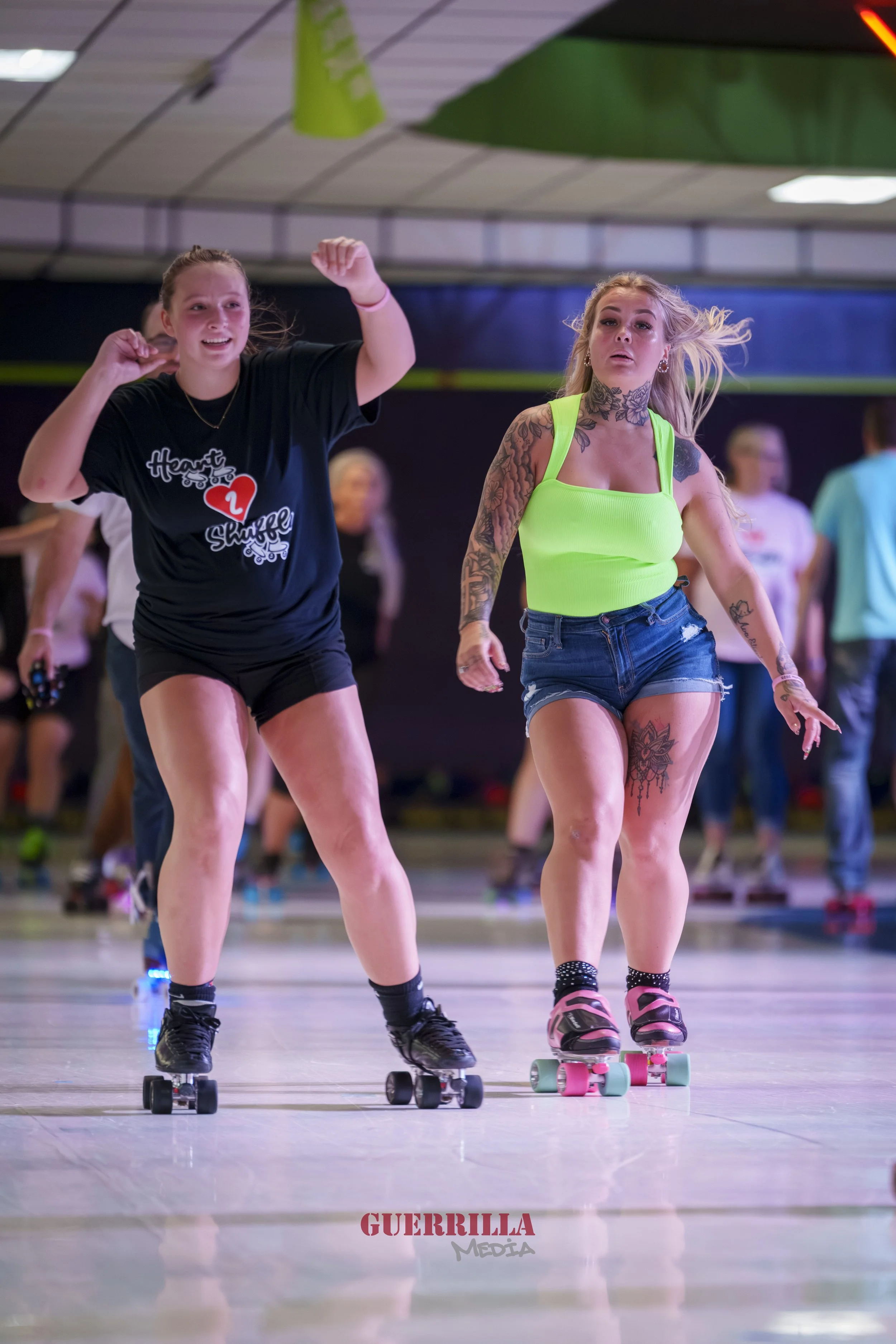 Two young women roller skating in an indoor skating rink, with other skaters in the background. One woman has tattoos and is wearing a bright green top and shorts, and the other woman is wearing a black t-shirt and shorts, both smiling and moving to 