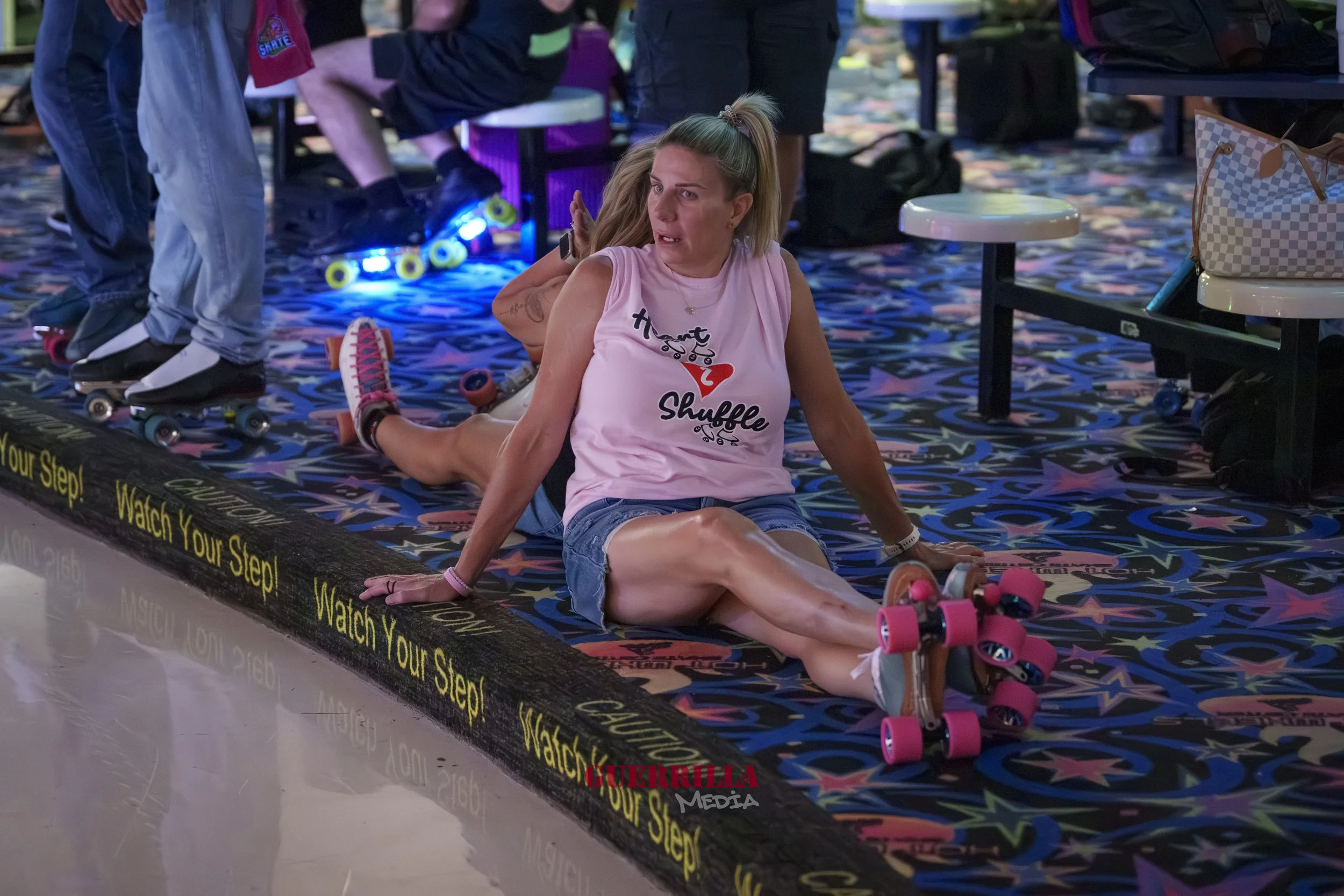 A woman is sitting on the floor at a roller skating rink, with her legs extended and hands supporting her. She has blonde hair tied in a high ponytail and is wearing a pink sleeveless shirt with the text "Heart Shuffle" and denim shorts. She is weari