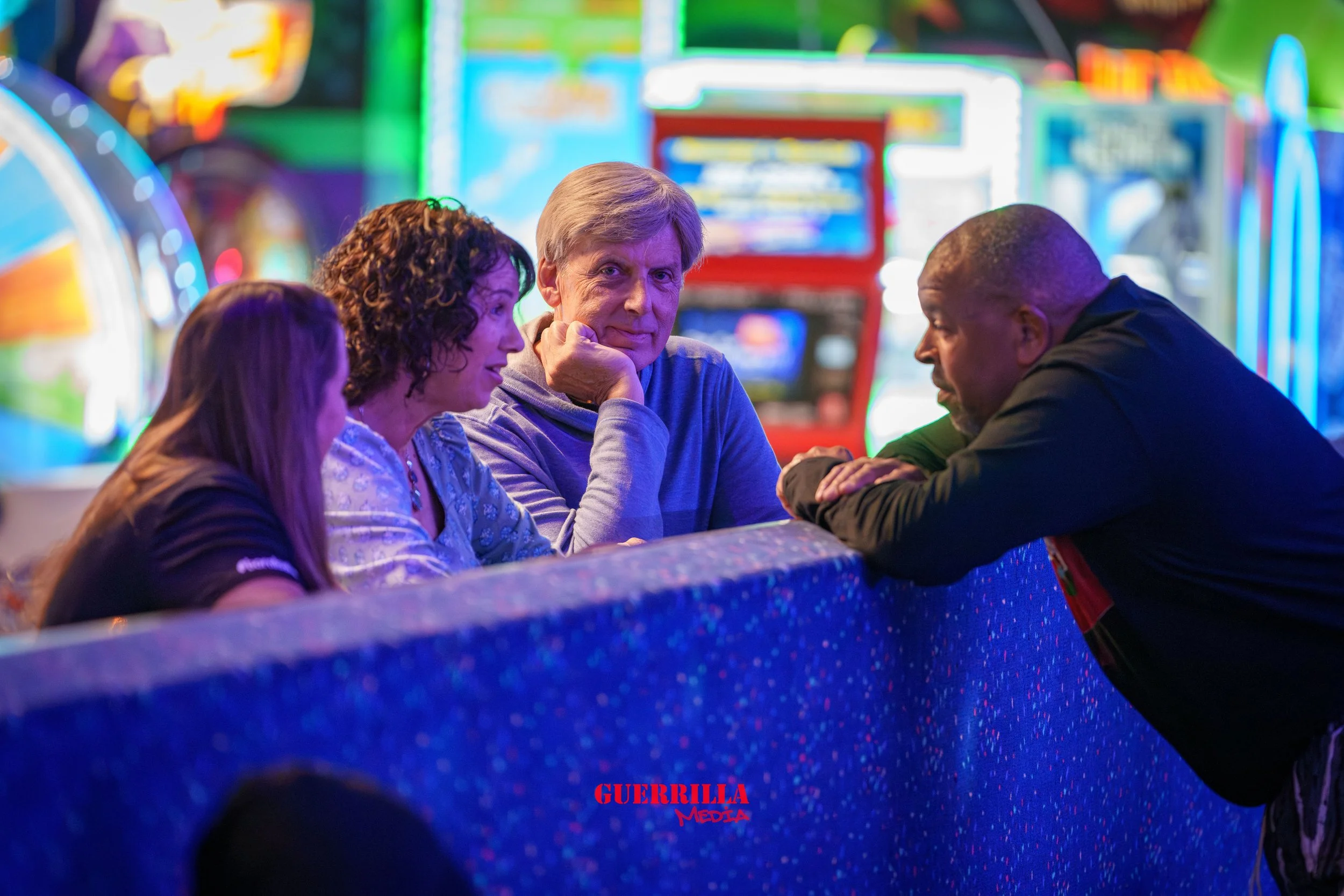 Four people sitting at a counter in front of bright arcade game machines in an arcade. They are engaged in conversation. One man leans on the counter and talks to a woman with curly hair. An older man with gray hair and a woman with long hair sit bes