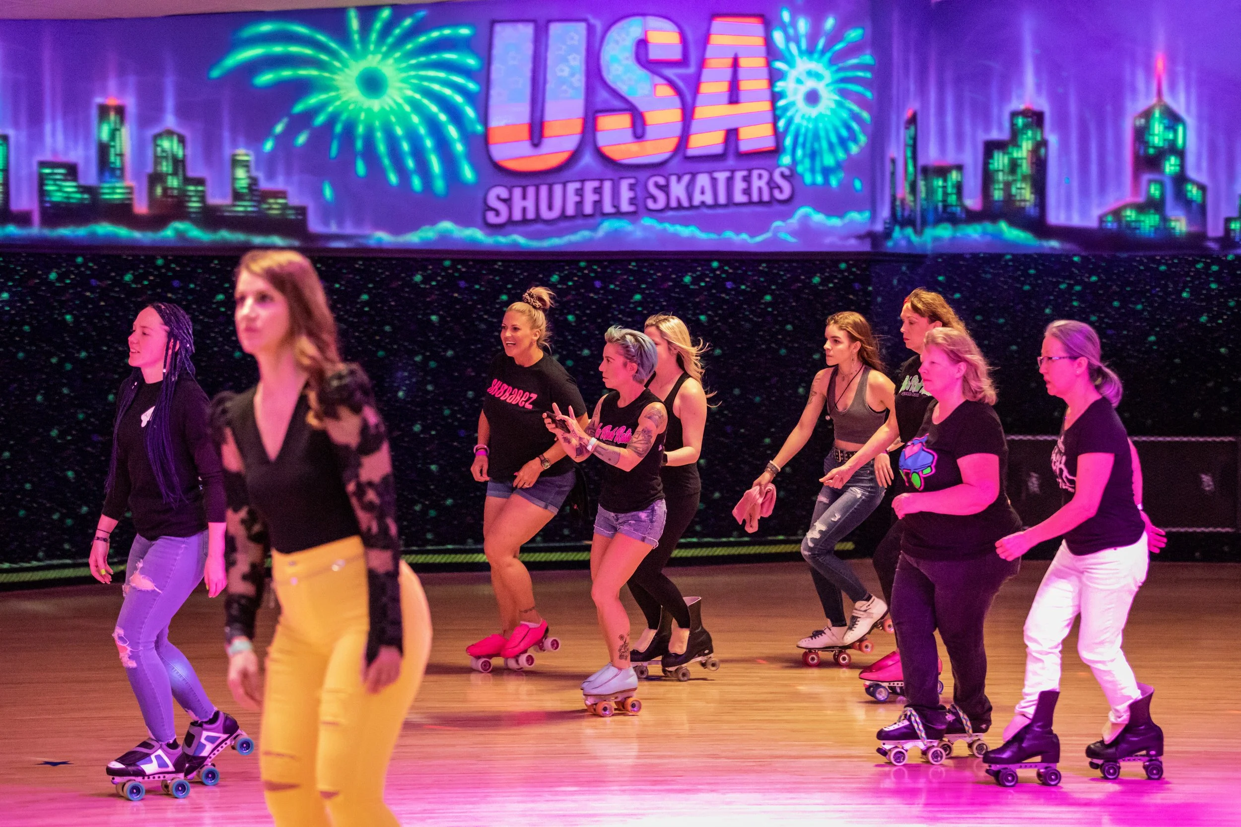 A group of women roller skating at an indoor rink with a neon sign that reads 'USSA Shuffle Skaters' and a city skyline with fireworks in the background.