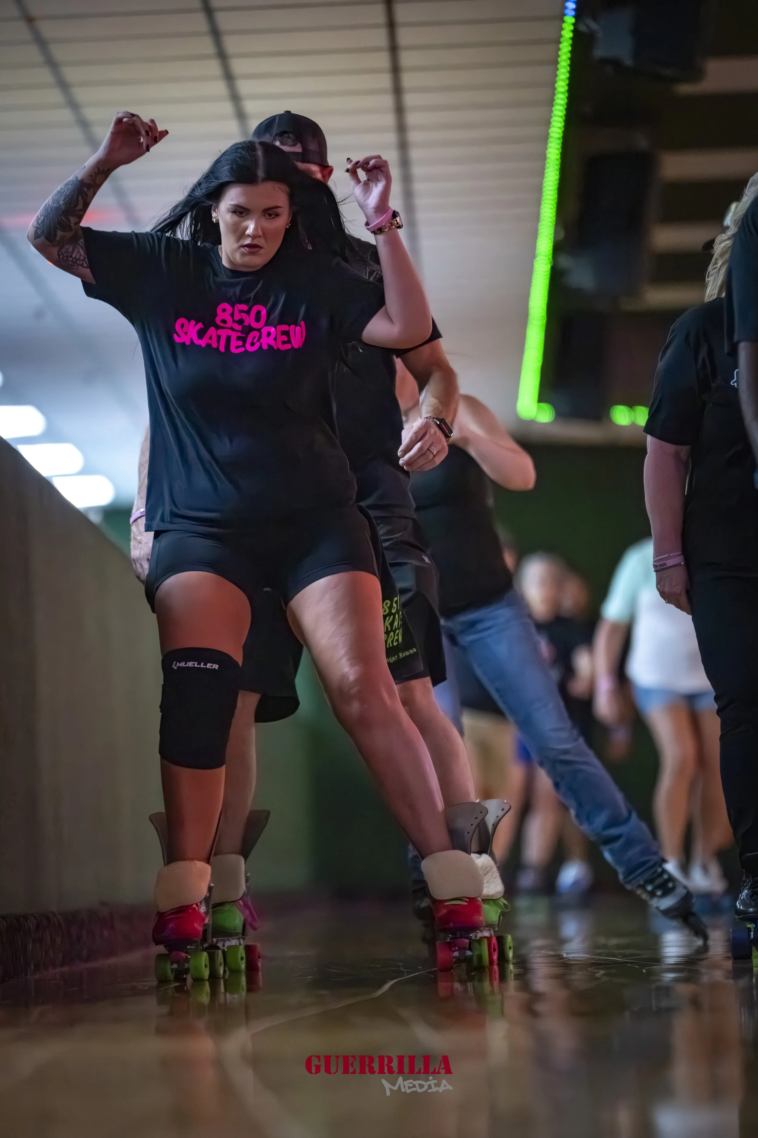 A woman roller skating indoors, wearing a black t-shirt that says '850 SKATECREW' in pink, black shorts, and black knee pads, with her arms raised and a serious expression.