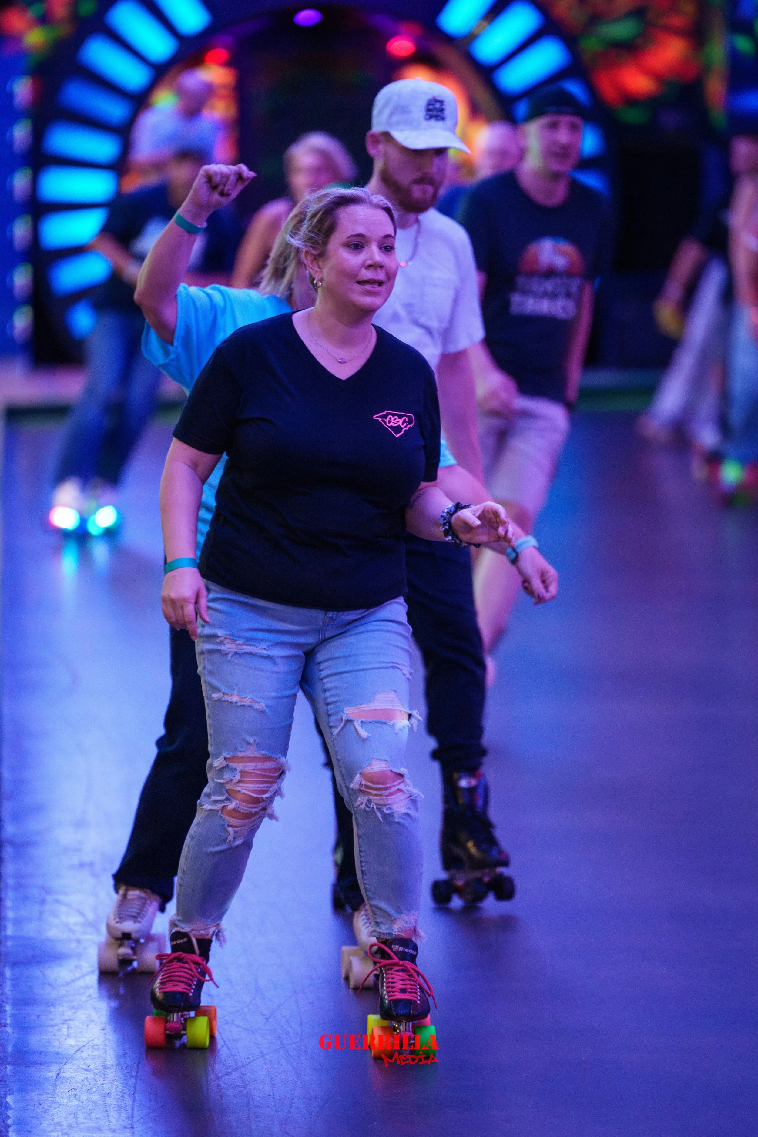 People roller skating indoors under colorful lights, with a woman in the front wearing a black t-shirt and ripped jeans leading the group.