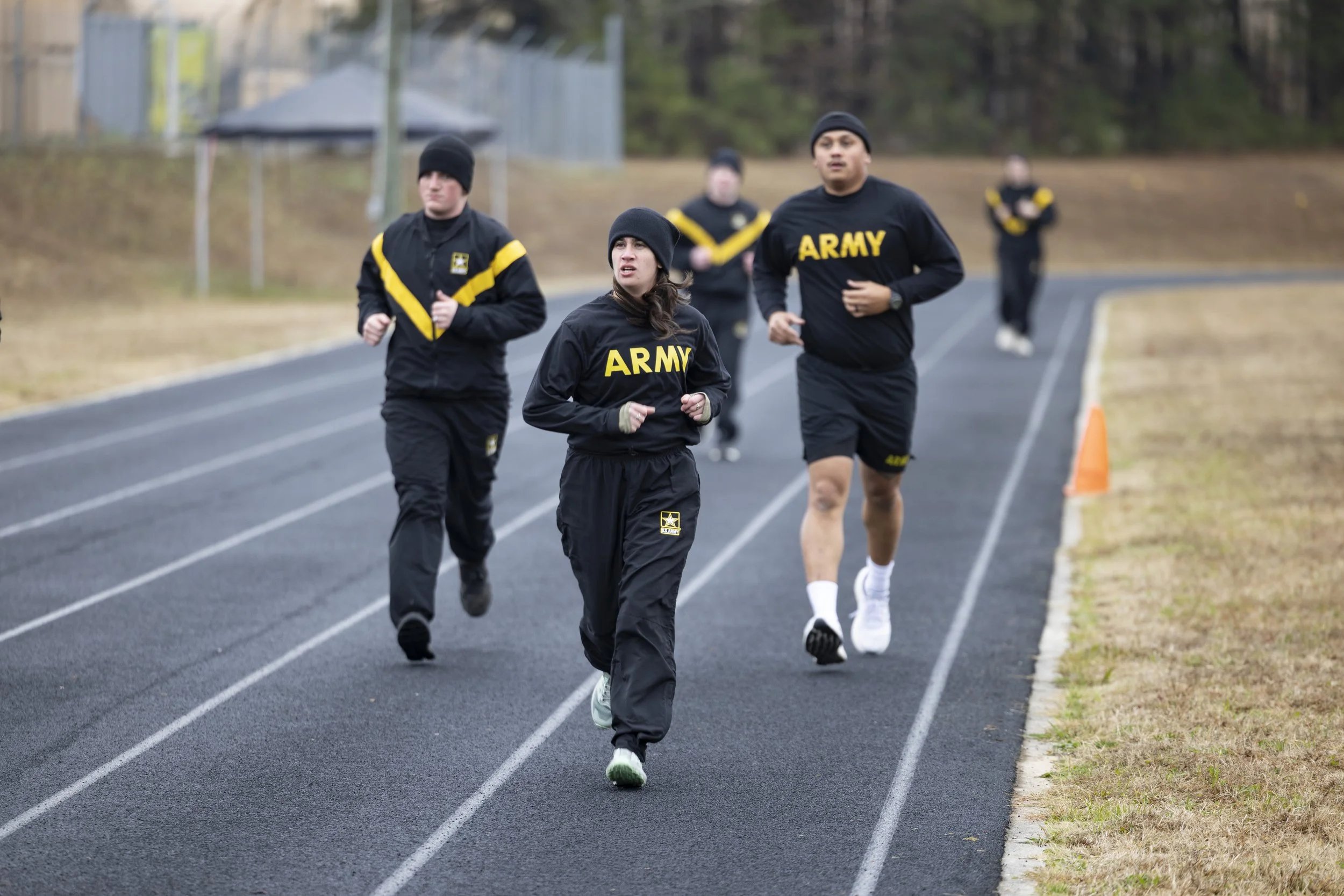 Group of Army personnel jogging on an outdoor track, wearing black athletic clothing with 'ARMY' written on their jackets, some in black beanies, in a natural setting with trees and a cone on the side of the track.