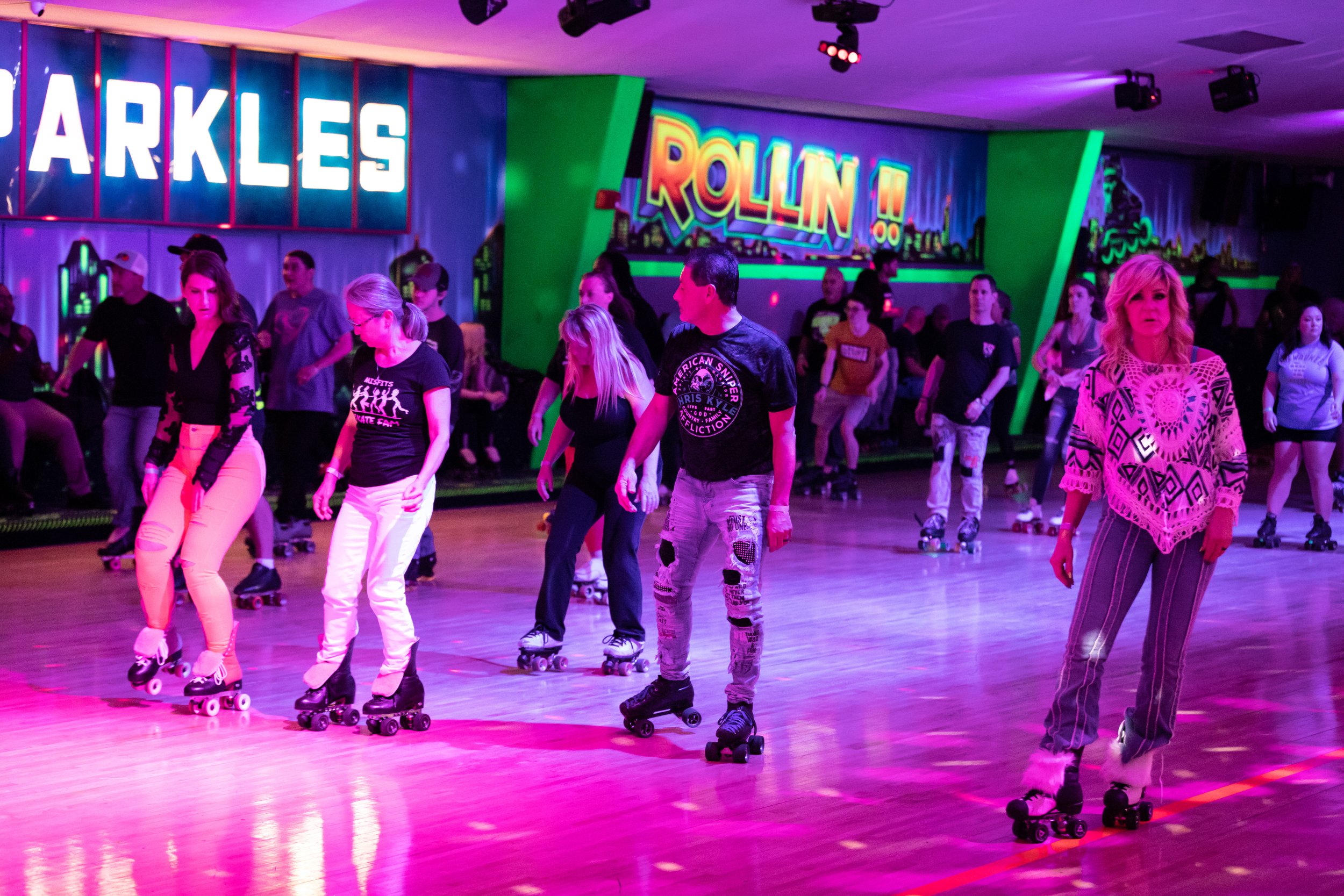People roller skating at an indoor rink with colorful neon and LED lights, a sign reading 'Sparkles' and a background sign that says 'Rollin'.