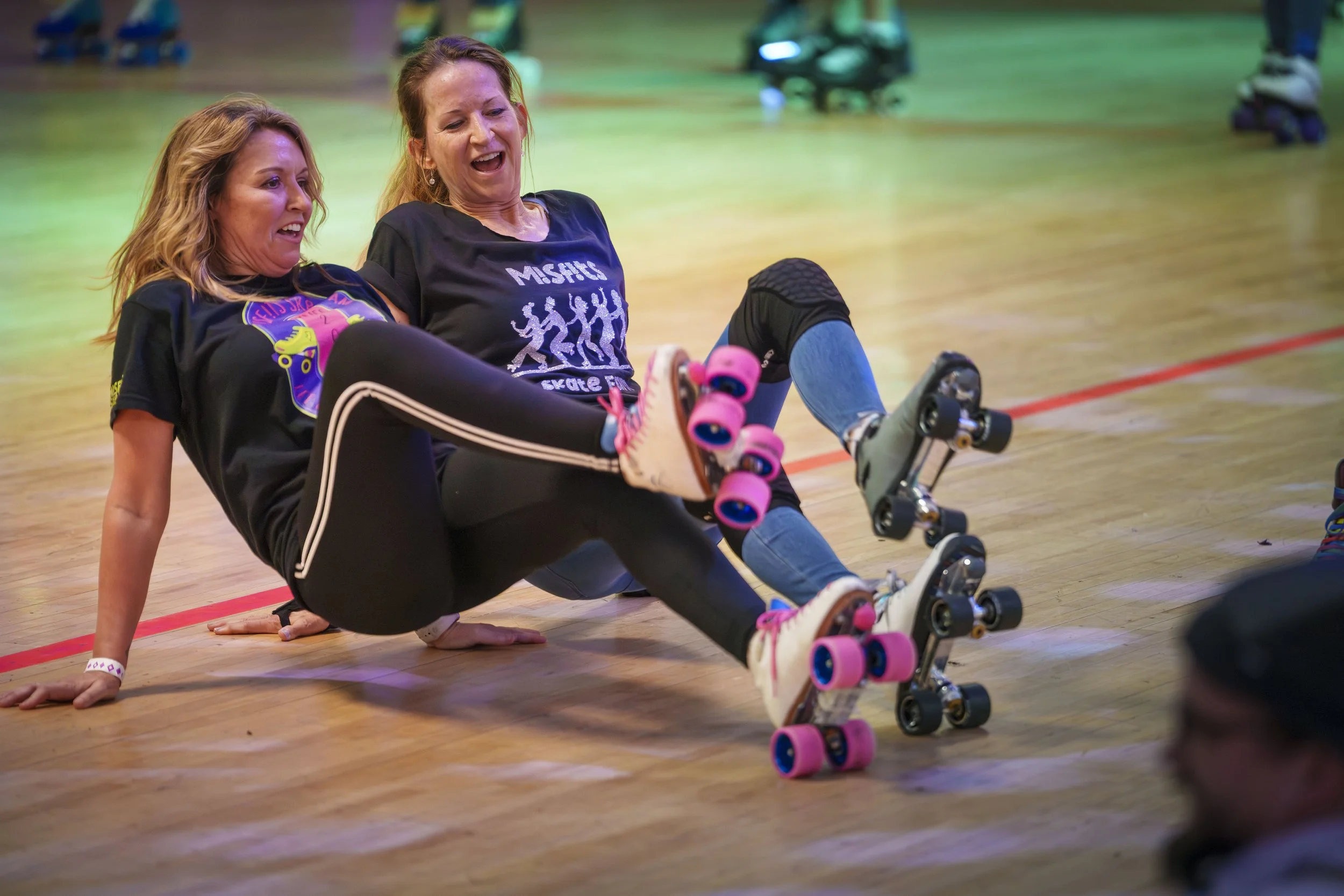 Two women in roller skates falling on a roller rink and laughing each other.