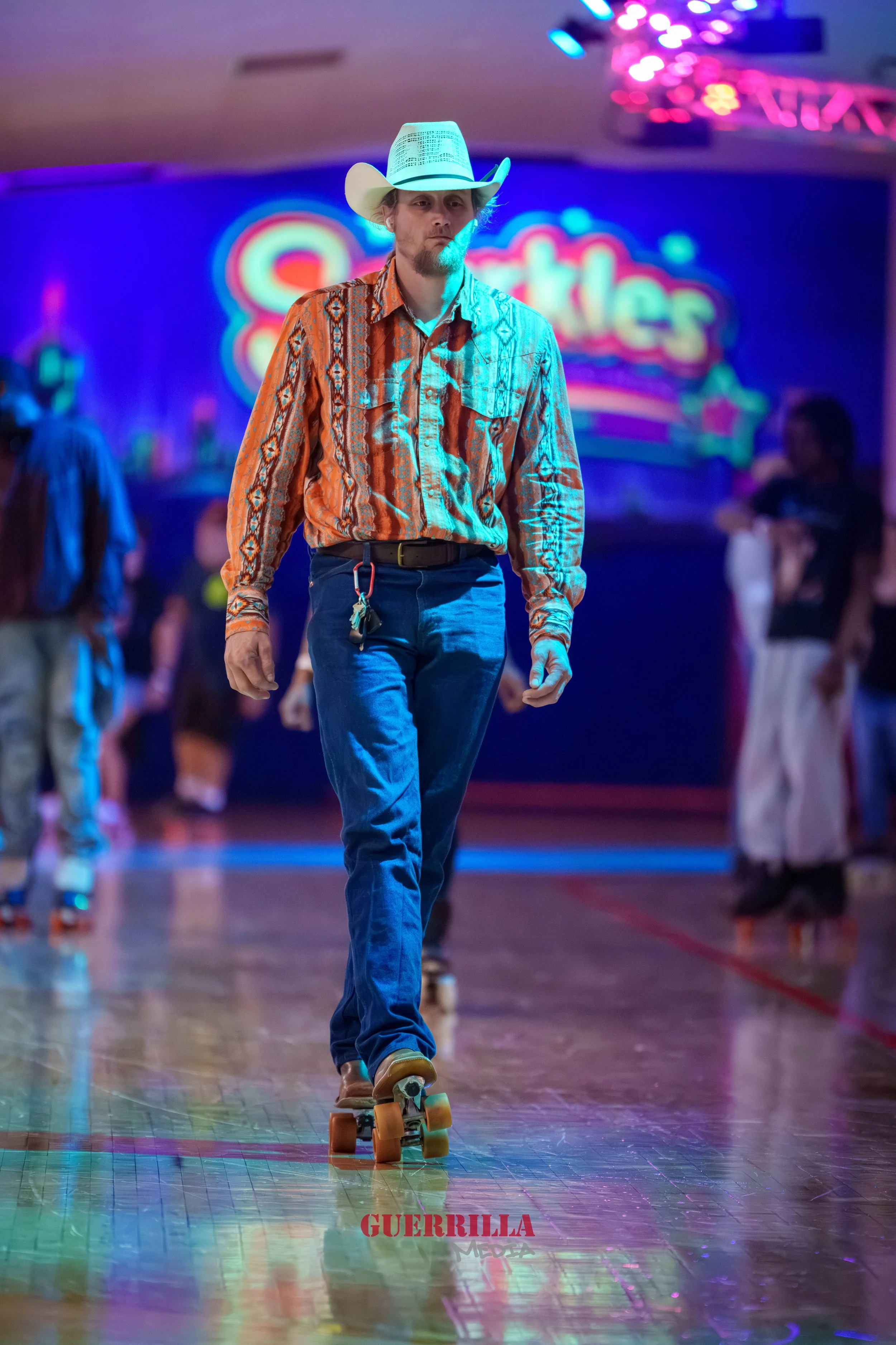 A person wearing a cowboy hat and jeans skateboarding on a wooden floor in a roller rink with colorful neon lights and a blurred crowd in the background.