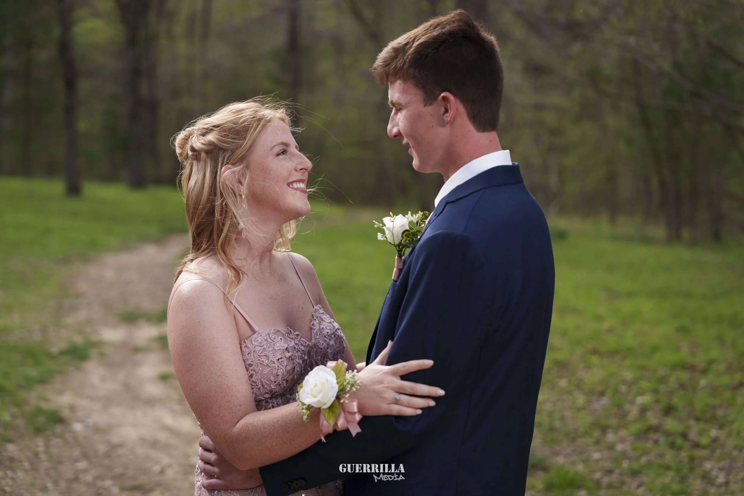 A young couple in wedding attire standing outdoors on a trail, looking at each other and smiling, surrounded by green trees and grass.