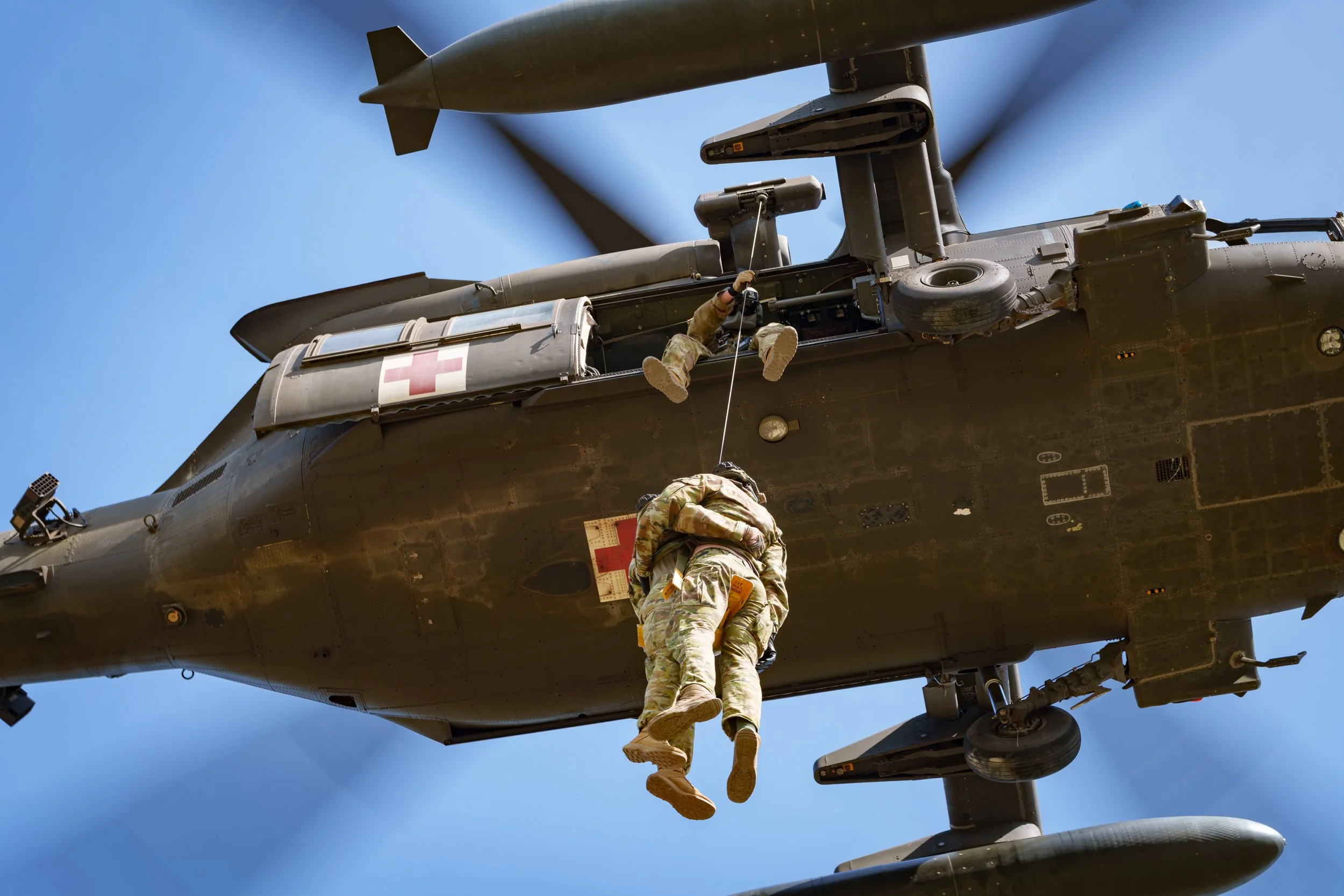 Two soldiers in camouflage uniforms rappelling from a military helicopter with medical crosses painted on it, in a clear blue sky.