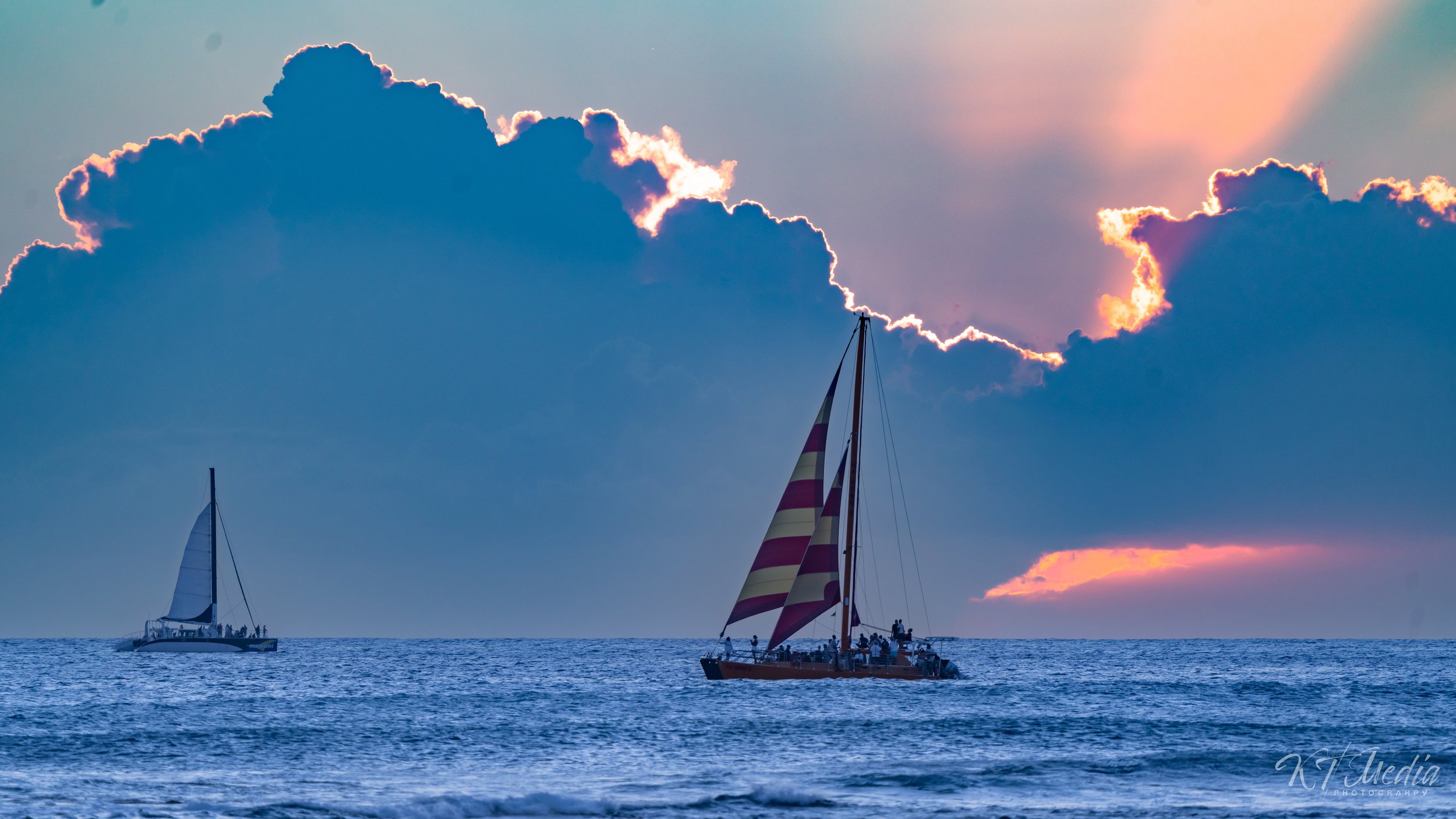 Two sailboats on the ocean during sunset with large clouds overhead, one with red and white striped sails, and the other with plain white sails.