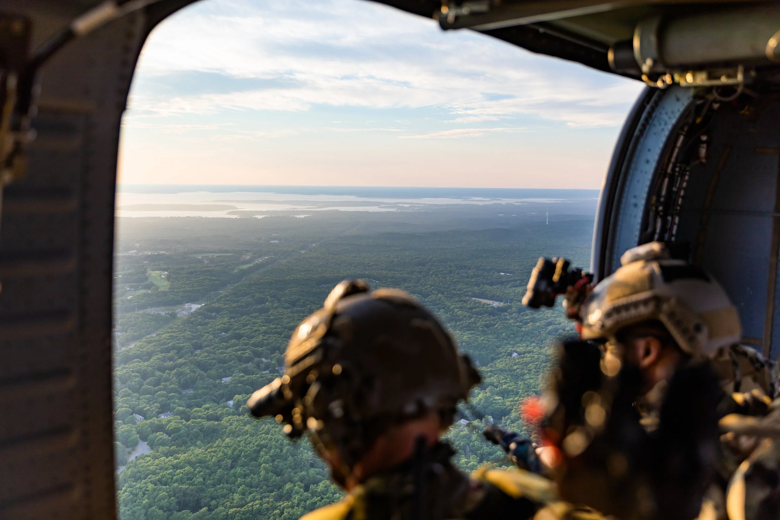 Two soldiers wearing helmets and tactical gear inside a military aircraft cockpit, flying over a green forested landscape with lakes and a distant horizon.