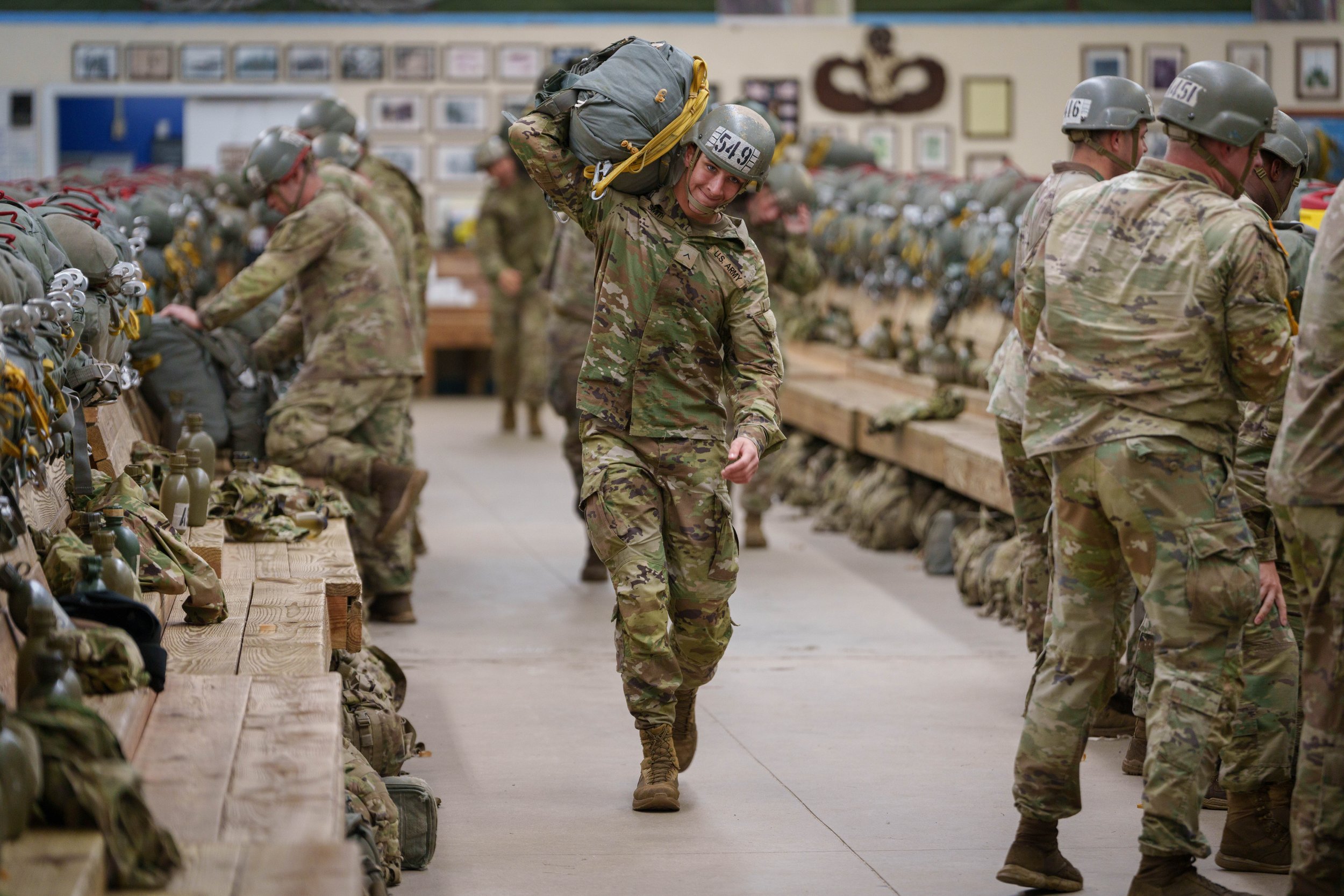 U.S. soldiers in camouflage uniforms and helmets shopping for military gear in an indoor store with rifles, backpacks, and other supplies on shelves.
