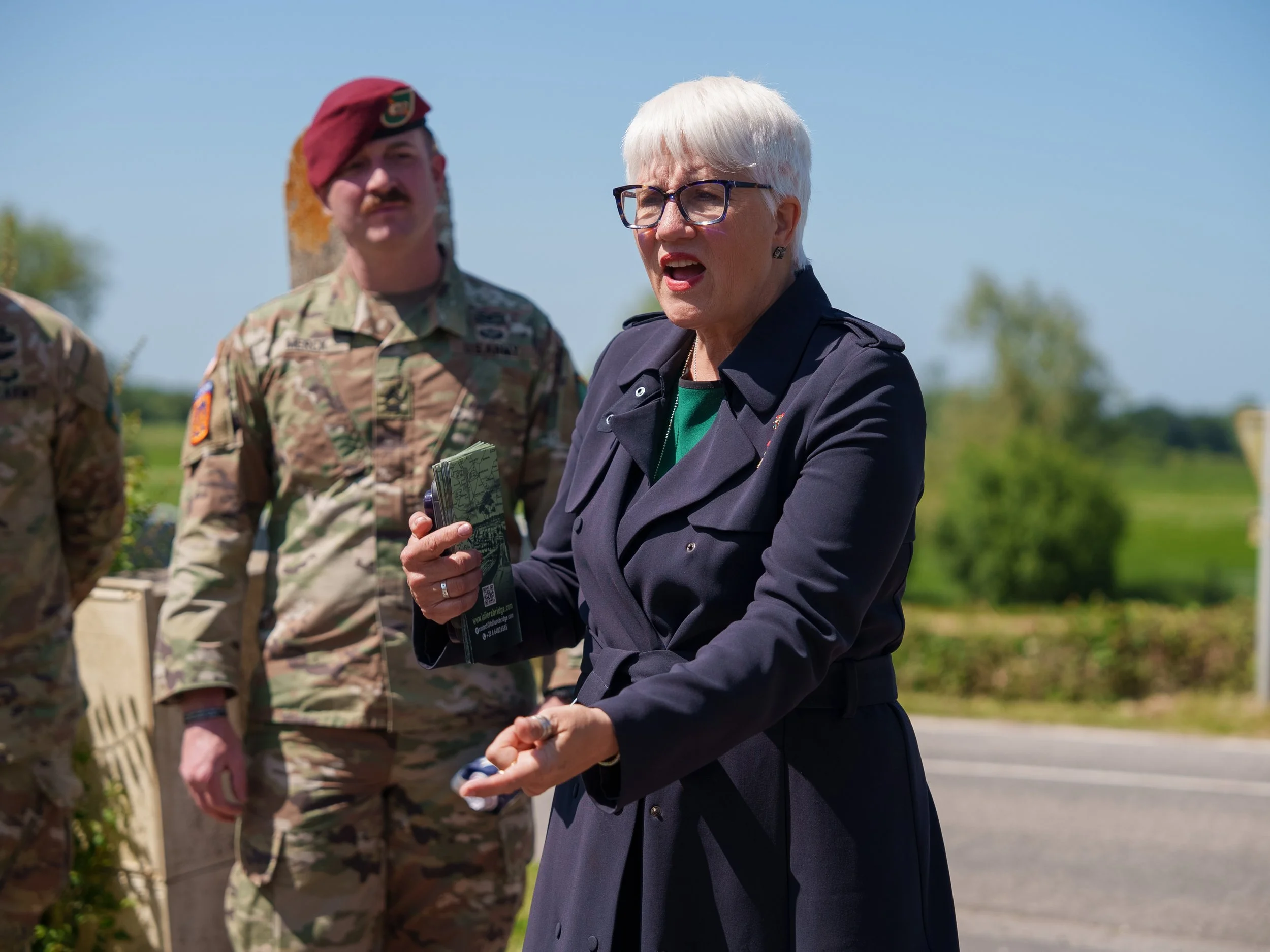 A woman with white hair, glasses, and a dark coat speaking outdoors, with military personnel in camouflage uniforms nearby under a clear blue sky.