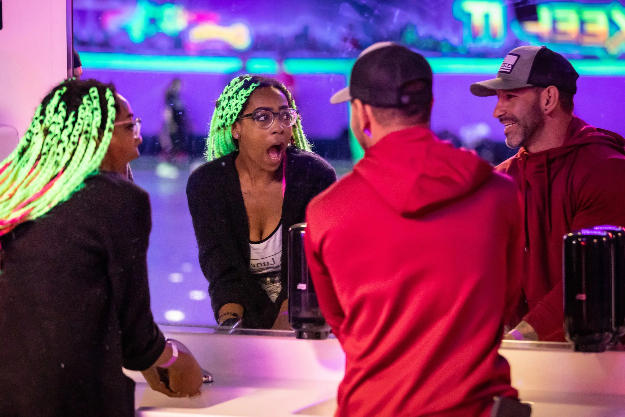 A group of five people at an indoor arcade or entertainment venue, with neon lights in the background. The woman in the center has bright green and black dreadlocks and appears surprised or shocked. The others are engaged in conversation, smiling or 