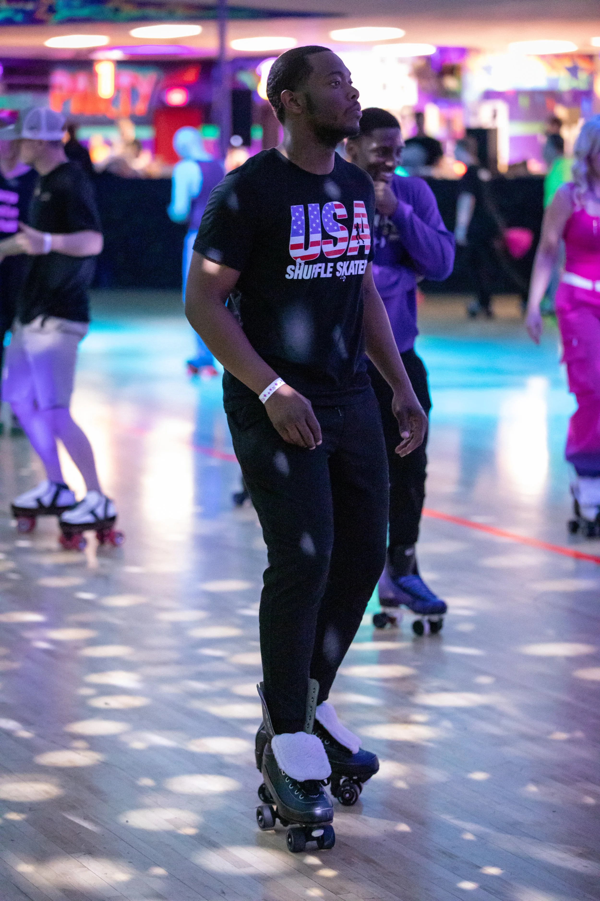 A young man roller skating at an indoor skating rink, wearing a black t-shirt with an American flag design and the words 'USA Shuffle Skater,' in a colorful, neon-lit environment with other skaters in the background.