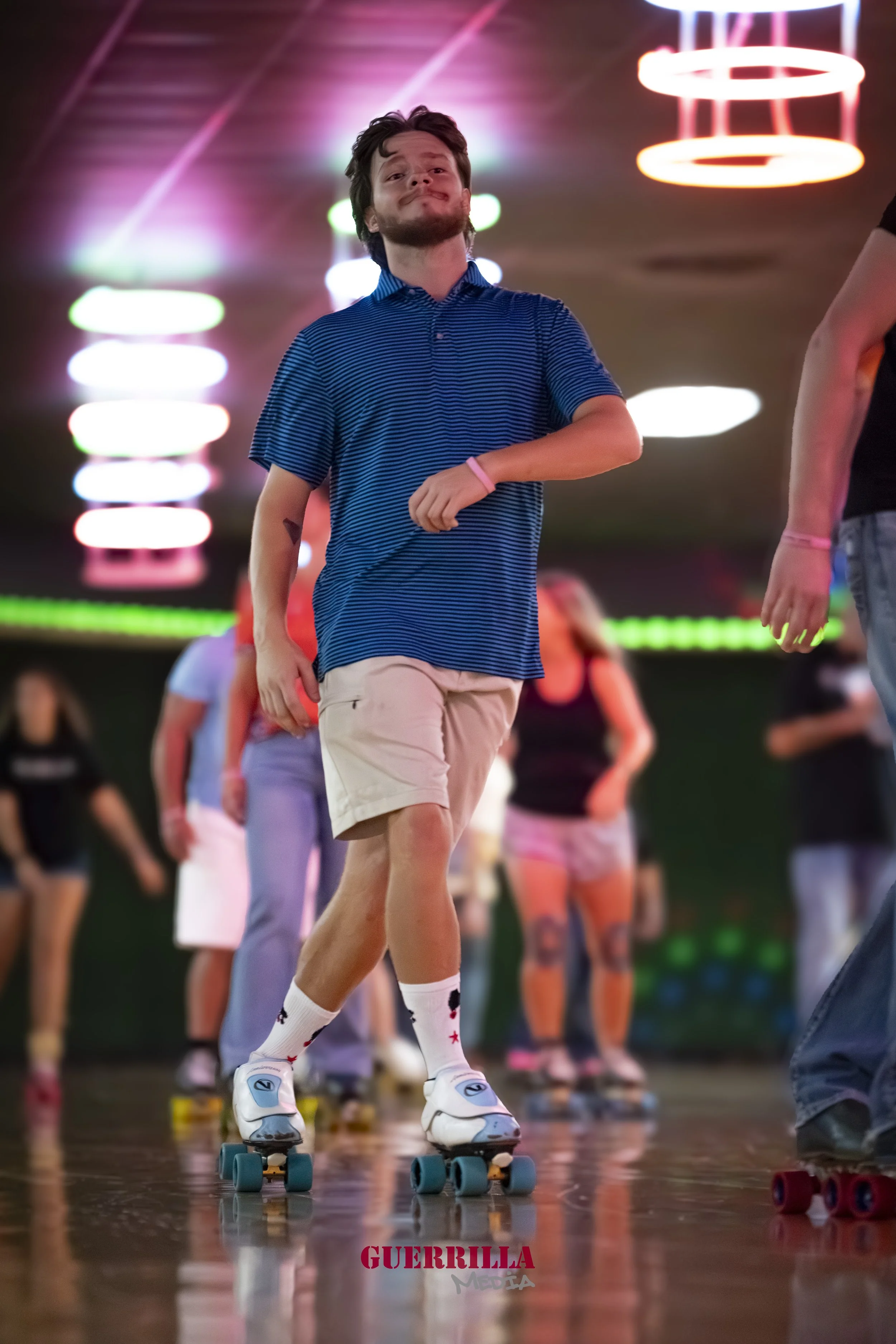 A young man roller skating in a roller rink with colorful neon lights overhead, wearing a blue striped polo shirt, beige shorts, and white roller skates.