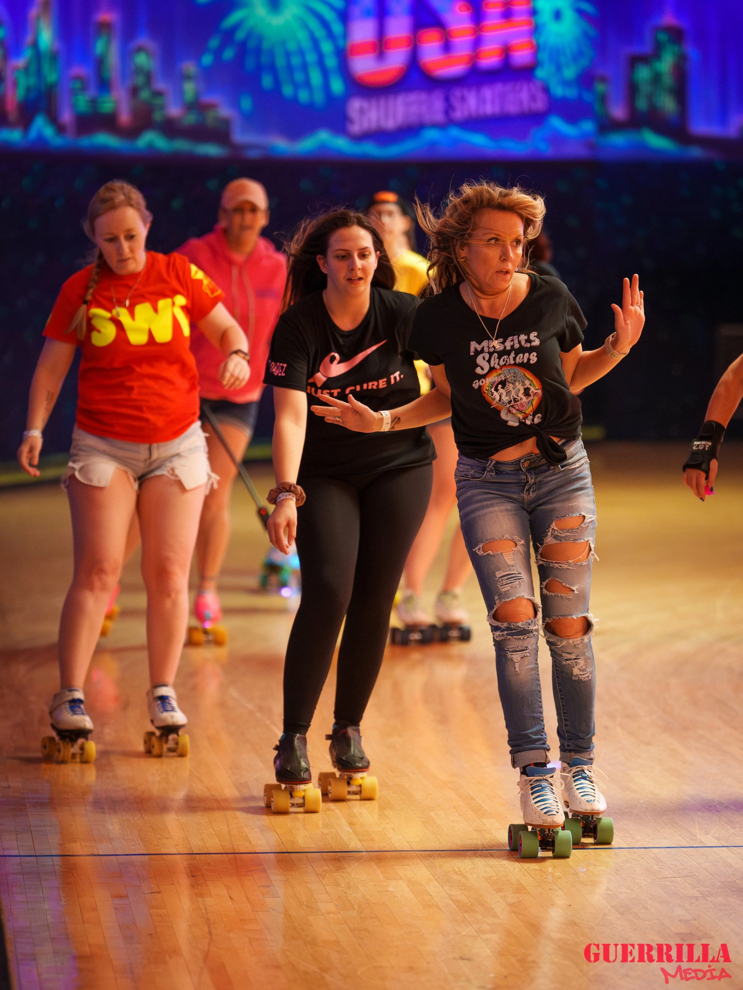 A group of people roller skating indoors on a wooden floor with colorful neon lights and a large digital screen in the background.