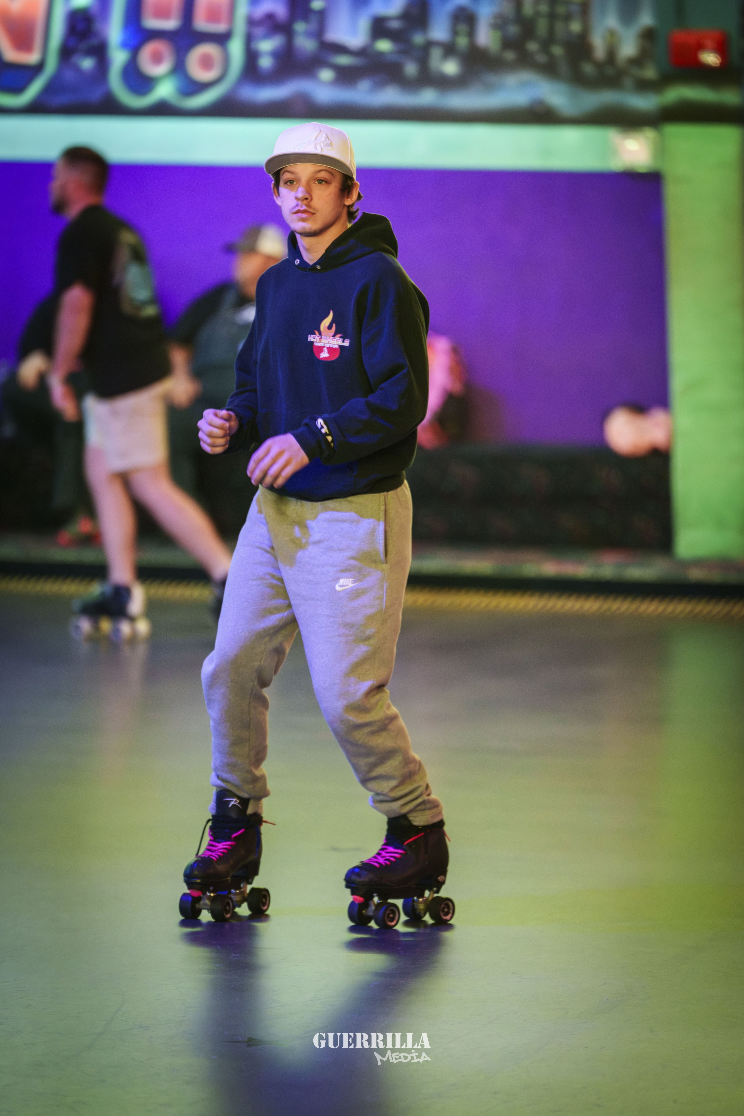 A young man roller skating indoors, wearing a black hoodie with a red logo, gray sweatpants, and a white baseball cap, in a colorful skating rink with other skaters in the background.