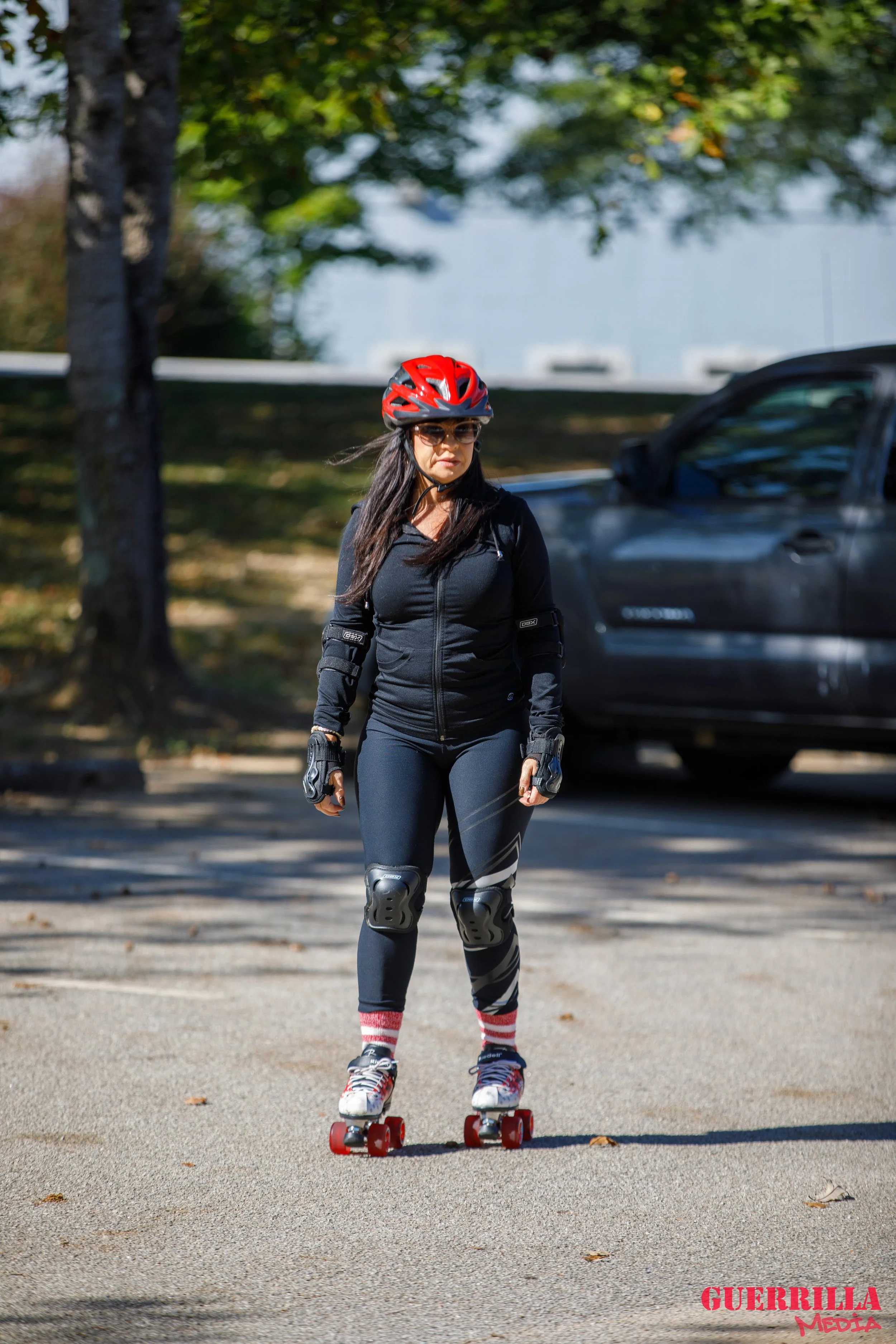 Woman wearing protective gear skateboarding on a paved surface outdoors with trees and a vehicle in the background.