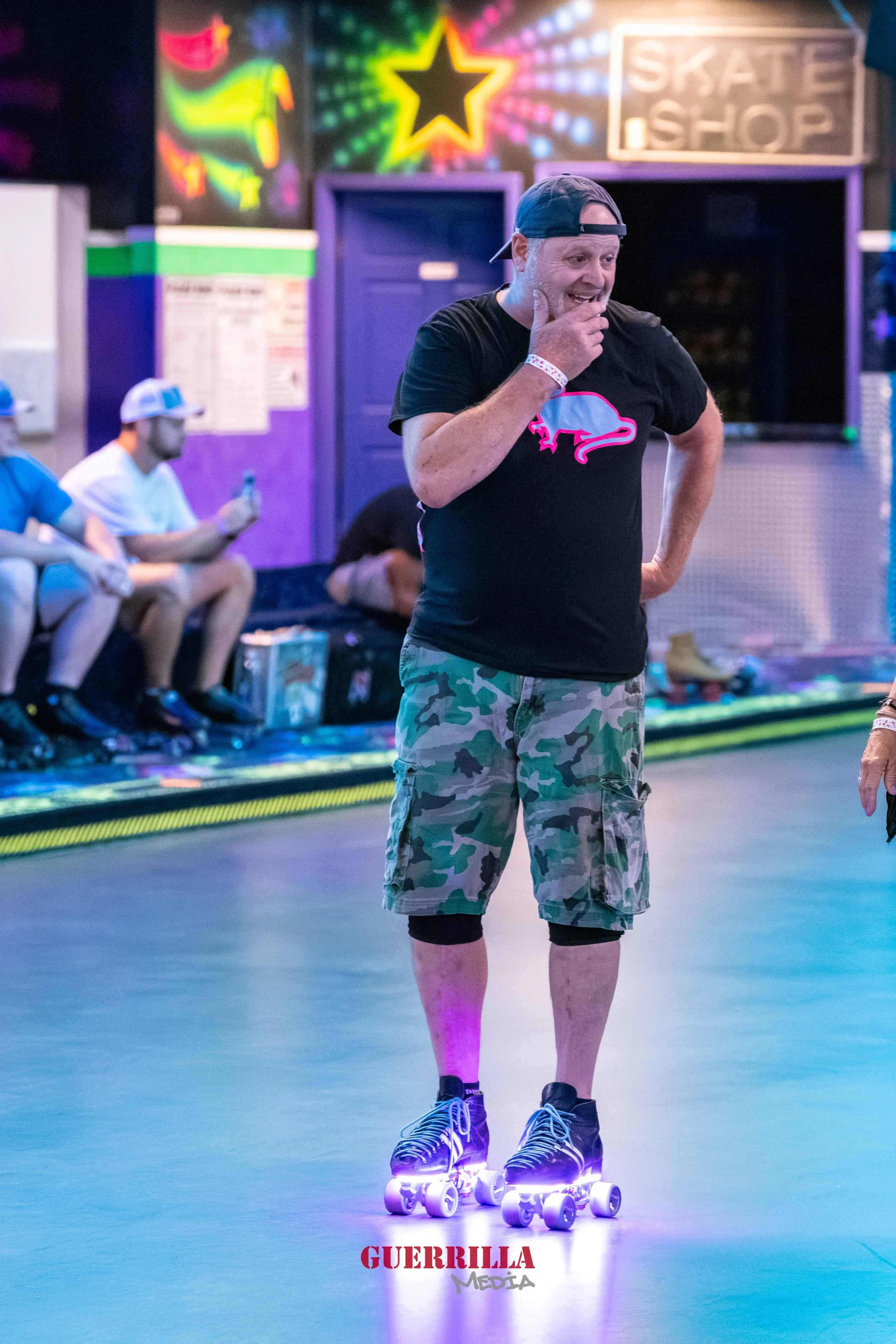 A man wearing a black t-shirt with a colorful buffalo logo, camo shorts, a backwards cap, and roller skates with glowing wheels, standing in an indoor roller skating rink with neon lights and a skate shop sign in the background.