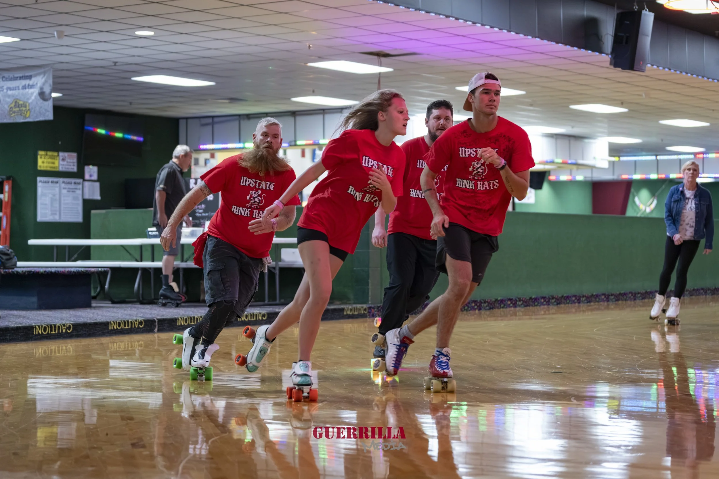Five people wearing red shirts roller skating indoors on a polished wooden floor, with rainbow-colored lights overhead.