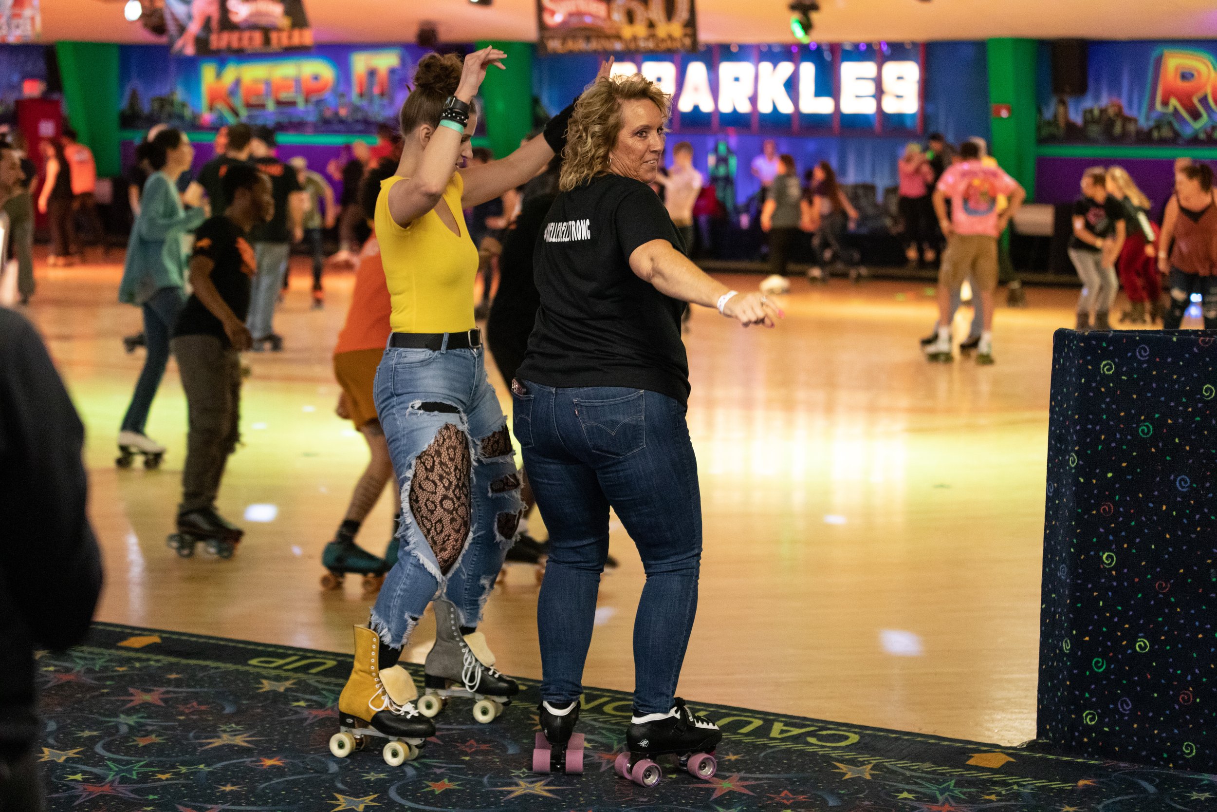 Two women roller skating at an indoor roller rink with a colorful, illuminated sign in the background that reads 'Keep It Sparkles.' One woman with curly hair is smiling and dancing, wearing a black shirt and jeans, while the other woman, with her ha