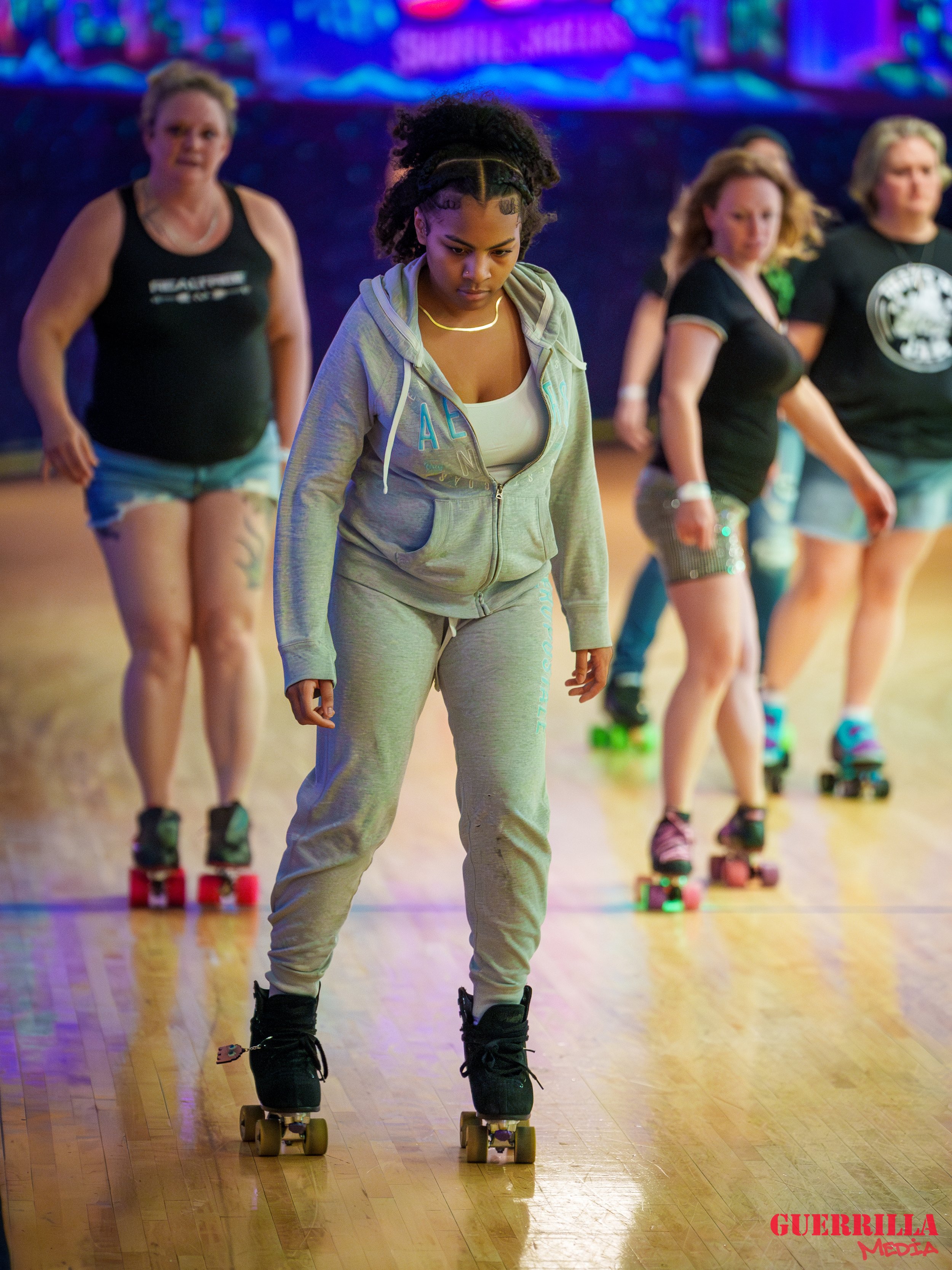 Group of women roller skating indoors, with one woman in the foreground wearing gray sweatpants and hoodie, looking down at her skates. The background is colorful and blurry.