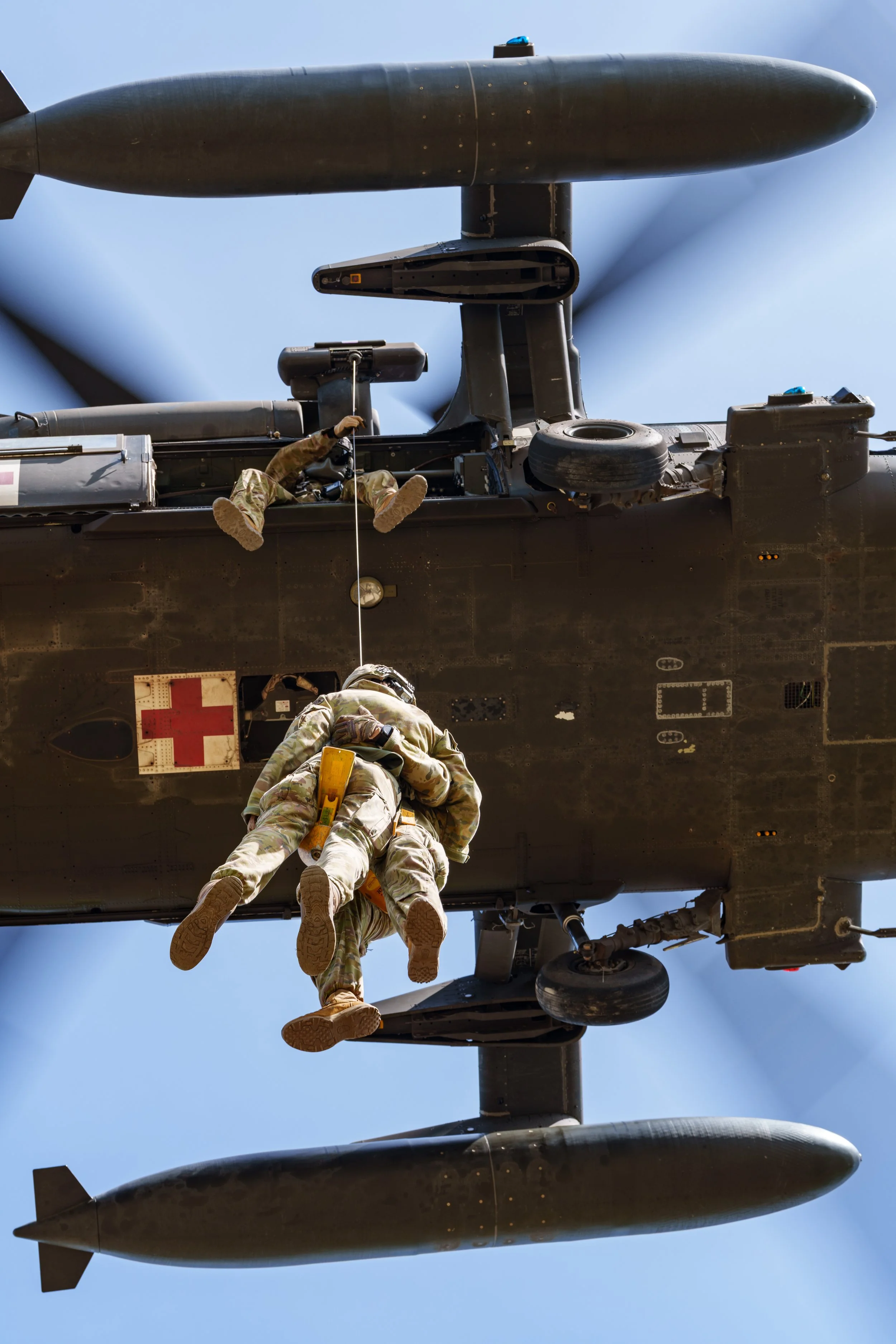 Military personnel evacuating a helicopter using a rope, with one person climbing up while another is hanging, under a cloudy blue sky.