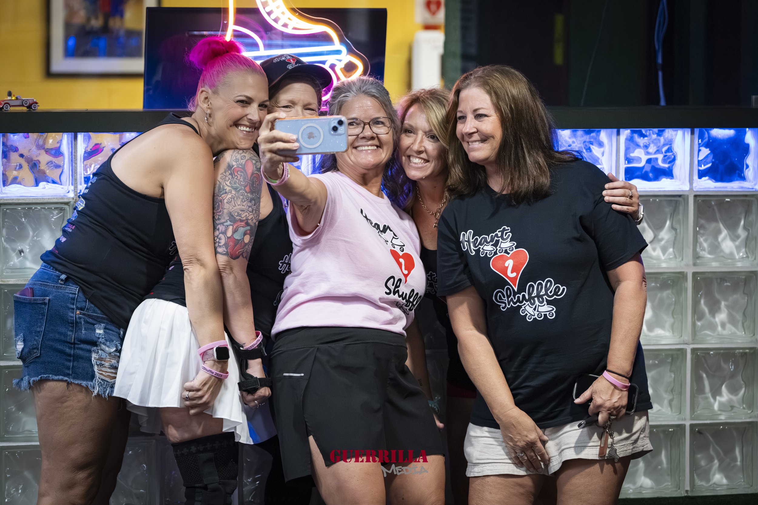 Five women smiling and taking a selfie, wearing matching t-shirts with a heart and roller skate design, in a colorful indoor setting with neon lights in the background.
