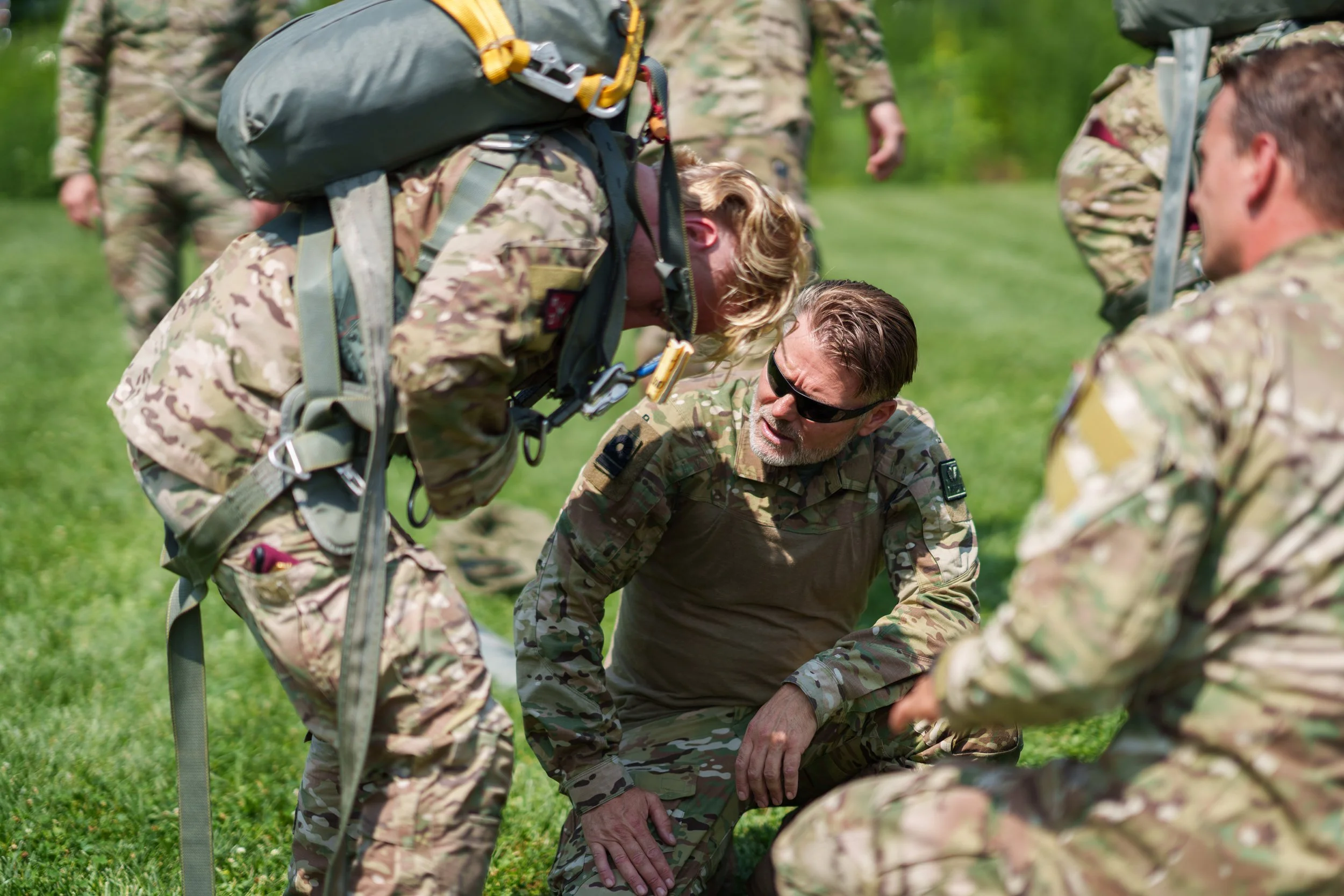Military personnel in camouflage uniforms assisting a kneeling soldier on the grass during training or a drill.