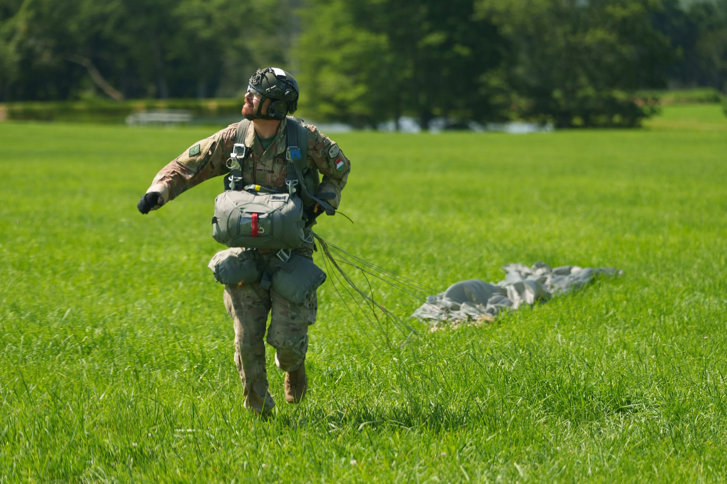 A soldier in camouflage uniform with a helmet and gear is running across a grassy field, pulling a parachute behind him on a sunny day.