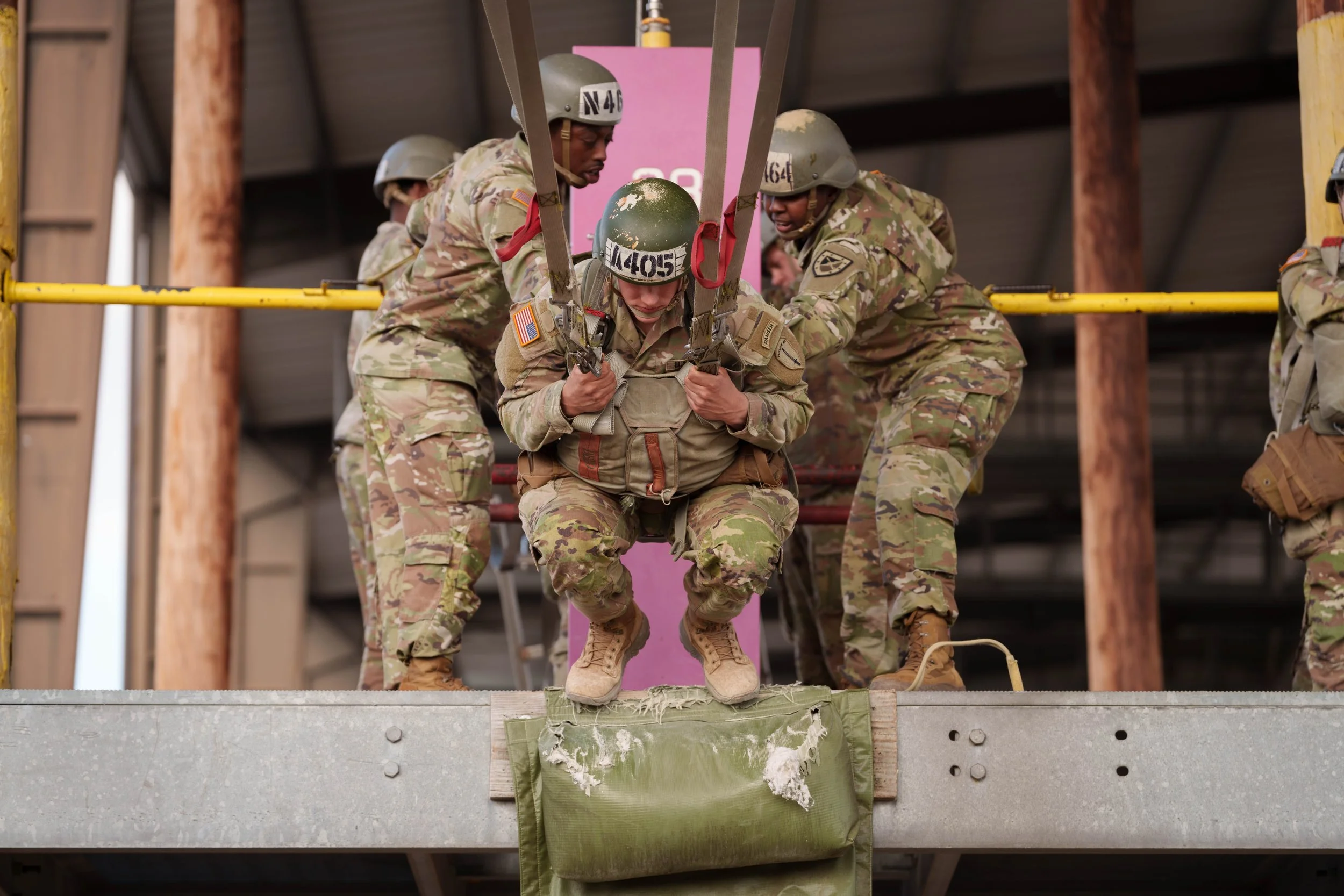 Military personnel participating in a training exercise, with one soldier harnessed and being guided by others during a rope rescue or rappel drill inside a building or training facility.