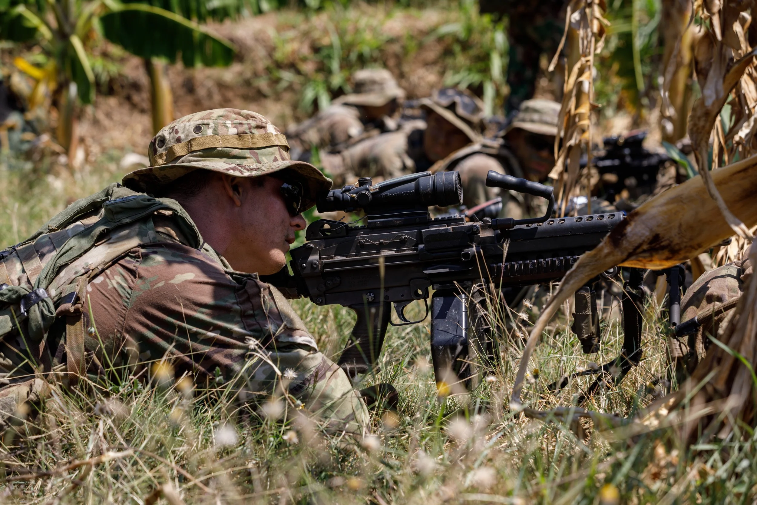 Military soldiers in camouflage uniforms lying on the ground in a grassy field, aiming a sniper rifle through binoculars, with several other soldiers in the background.